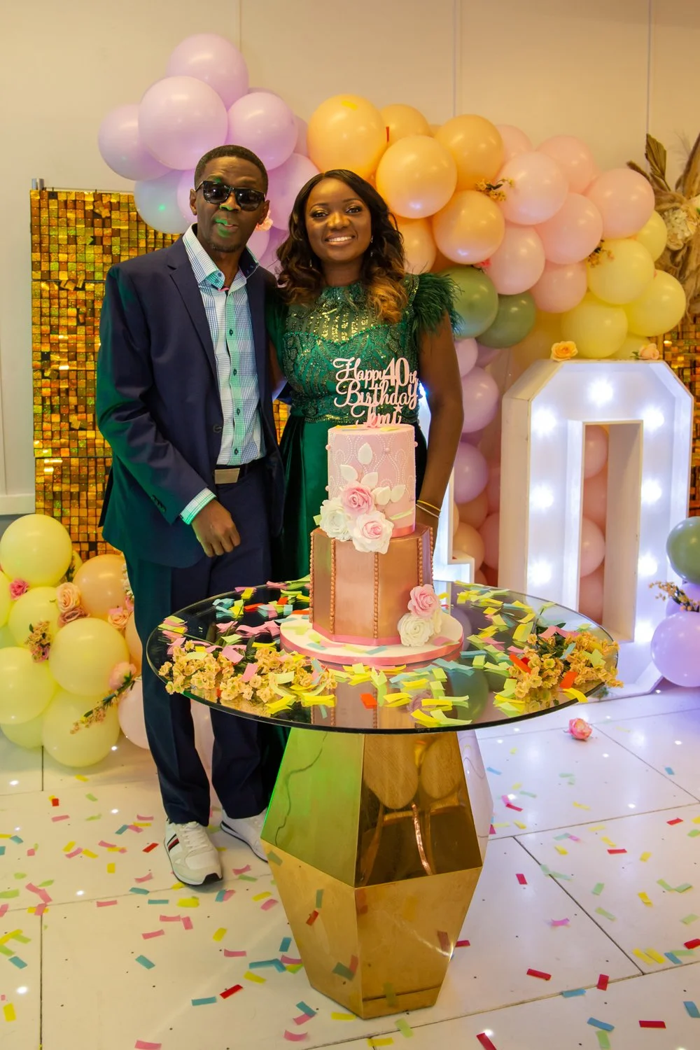 A woman celebrating her 40th birthday with a man in front of a balloon backdrop, with a decorated cake on a reflective table surrounded by confetti and flowers.