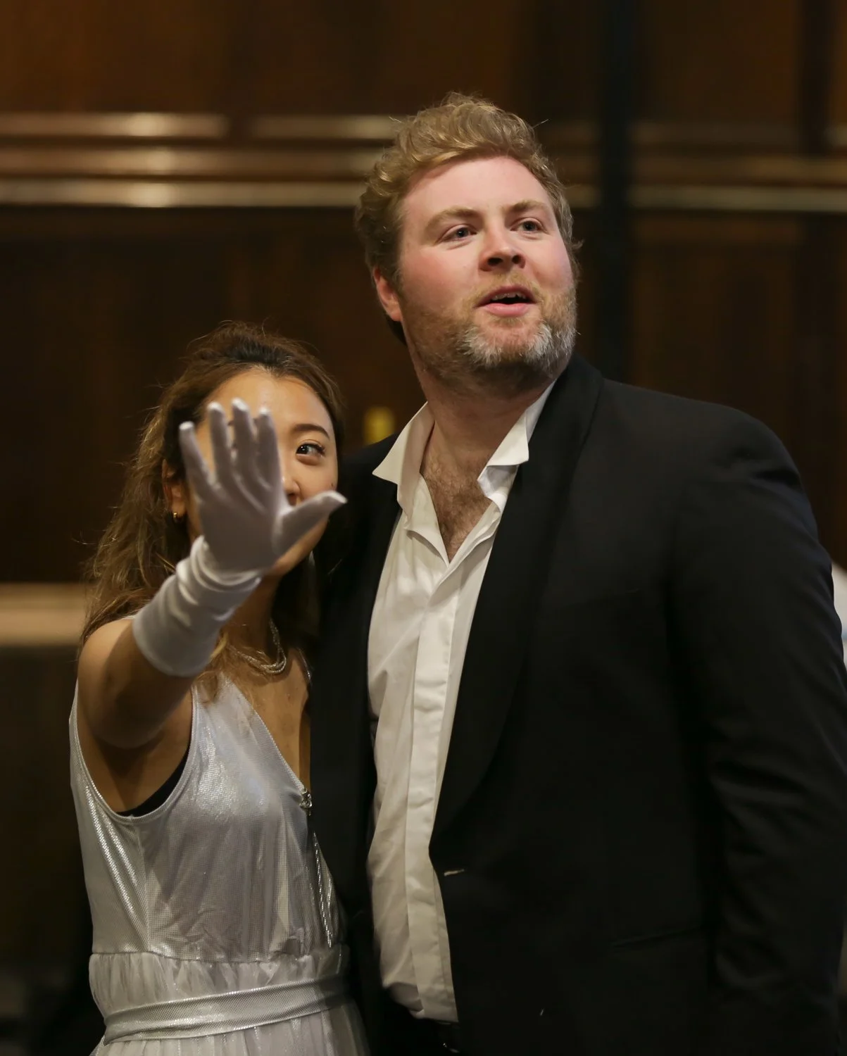 A man and a woman dressed in formal attire, with the woman waving at the camera, in an indoor setting with wooden paneling.