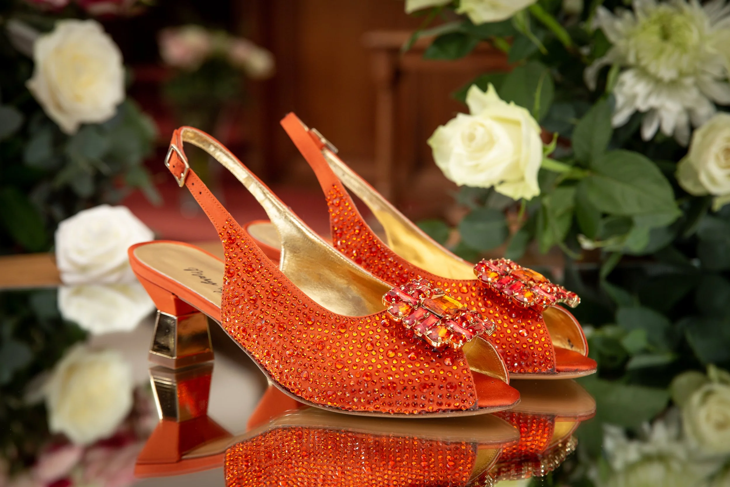 Orange high-heeled shoes decorated with rhinestones and large jeweled embellishments on the toes, placed on a reflective surface with white roses and green foliage in the background.