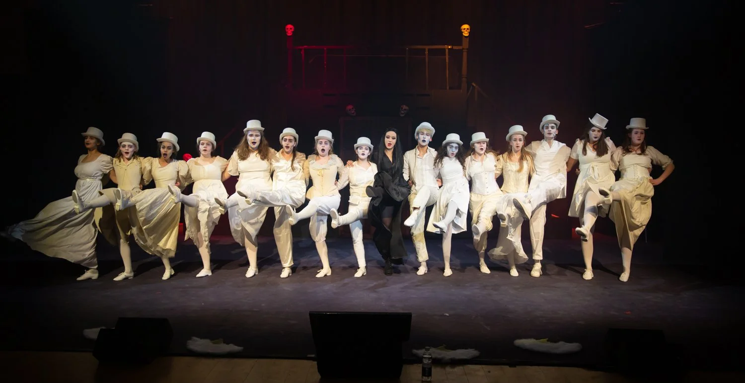 A theatrical performance featuring 14 performers dressed in white costumes and bowler hats, with a dark background and colorful lights.
