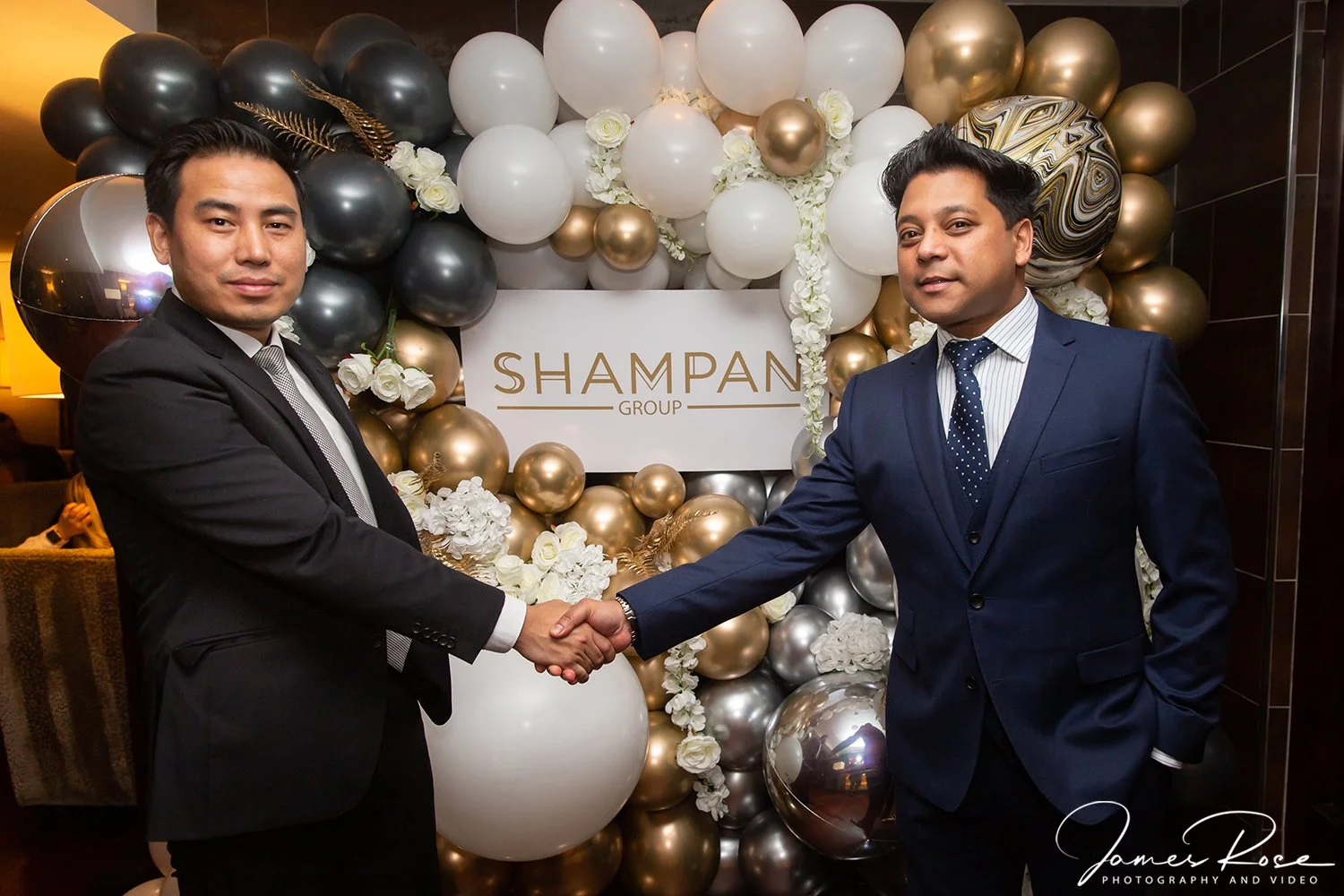 Two men in formal suits shaking hands at an event with a backdrop of black, white, gold, and marbled balloons and a sign reading 'Shampan Group'
