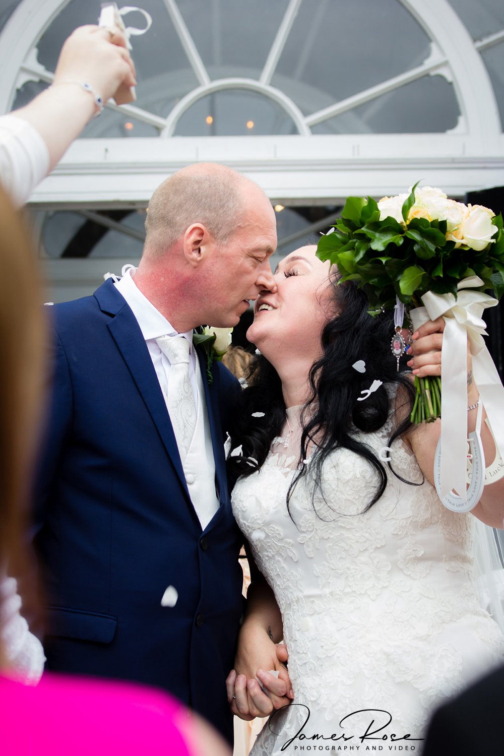 A bride and groom sharing a kiss during their wedding, with the bride holding a bouquet of white flowers, surrounded by guests.