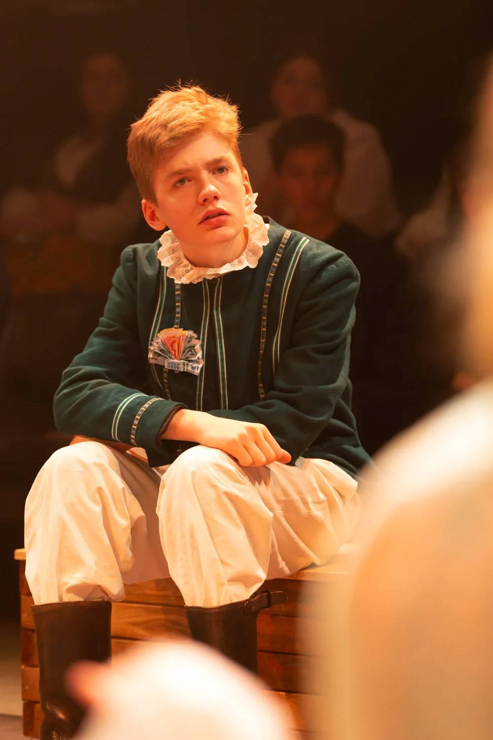 Young person with red hair in period costume, sitting on a wooden platform, with a serious expression, in a theatrical or stage setting with other blurred people in the background.