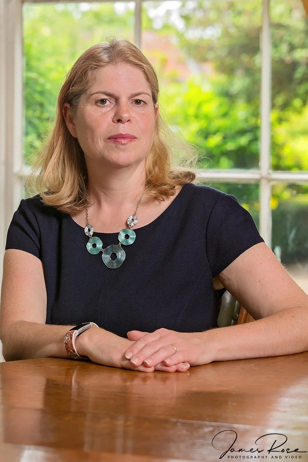A woman with blonde hair wearing a black shirt, a circular turquoise and silver necklace, sitting at a wooden table with a window showing green outdoor scenery in the background.