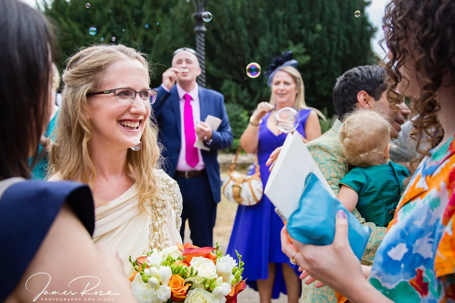 A woman holding a bouquet of flowers smiling at a gathering of people outdoors, with others in formal and colorful attire, including a woman in blue and a man in a dark suit with a pink tie, surrounded by bubbles.