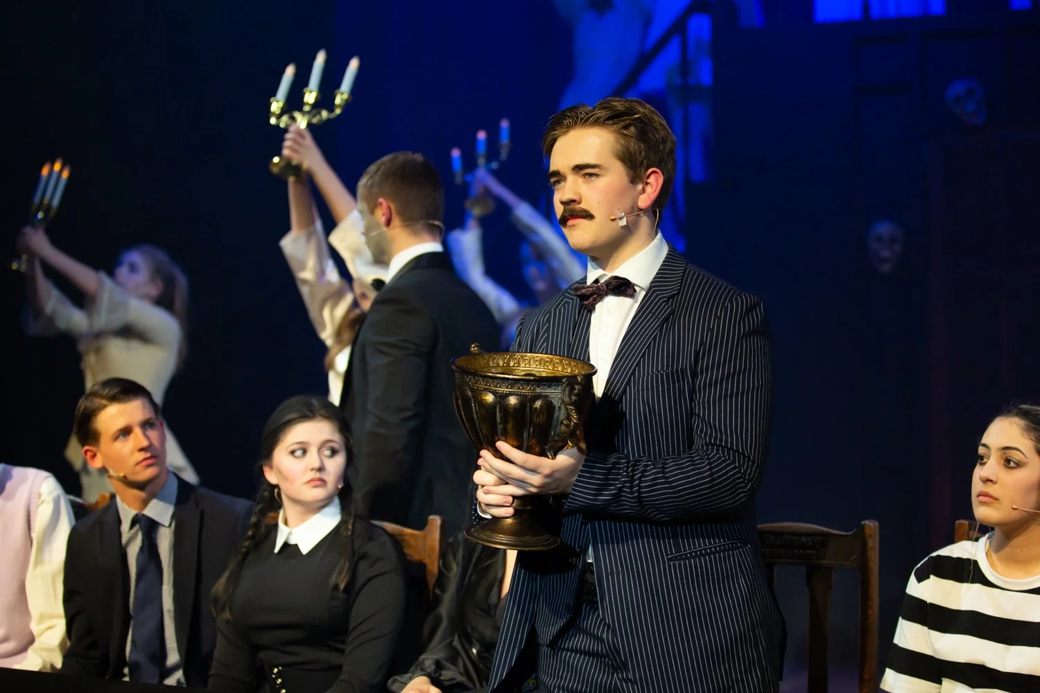 Man in pinstripe suit with mustache holding a gold bowl on stage, with seated and standing people in period costumes, some holding candelabra, during a theatrical performance.