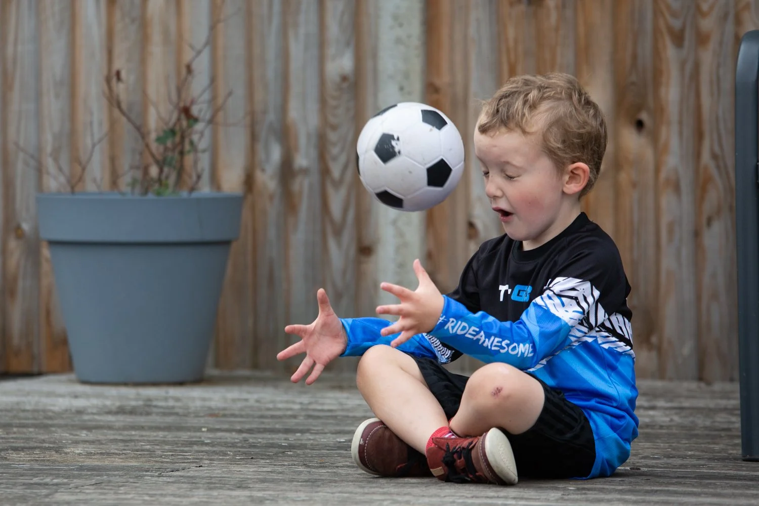 Young boy sitting on a wooden deck outdoors, playing with a soccer ball.
