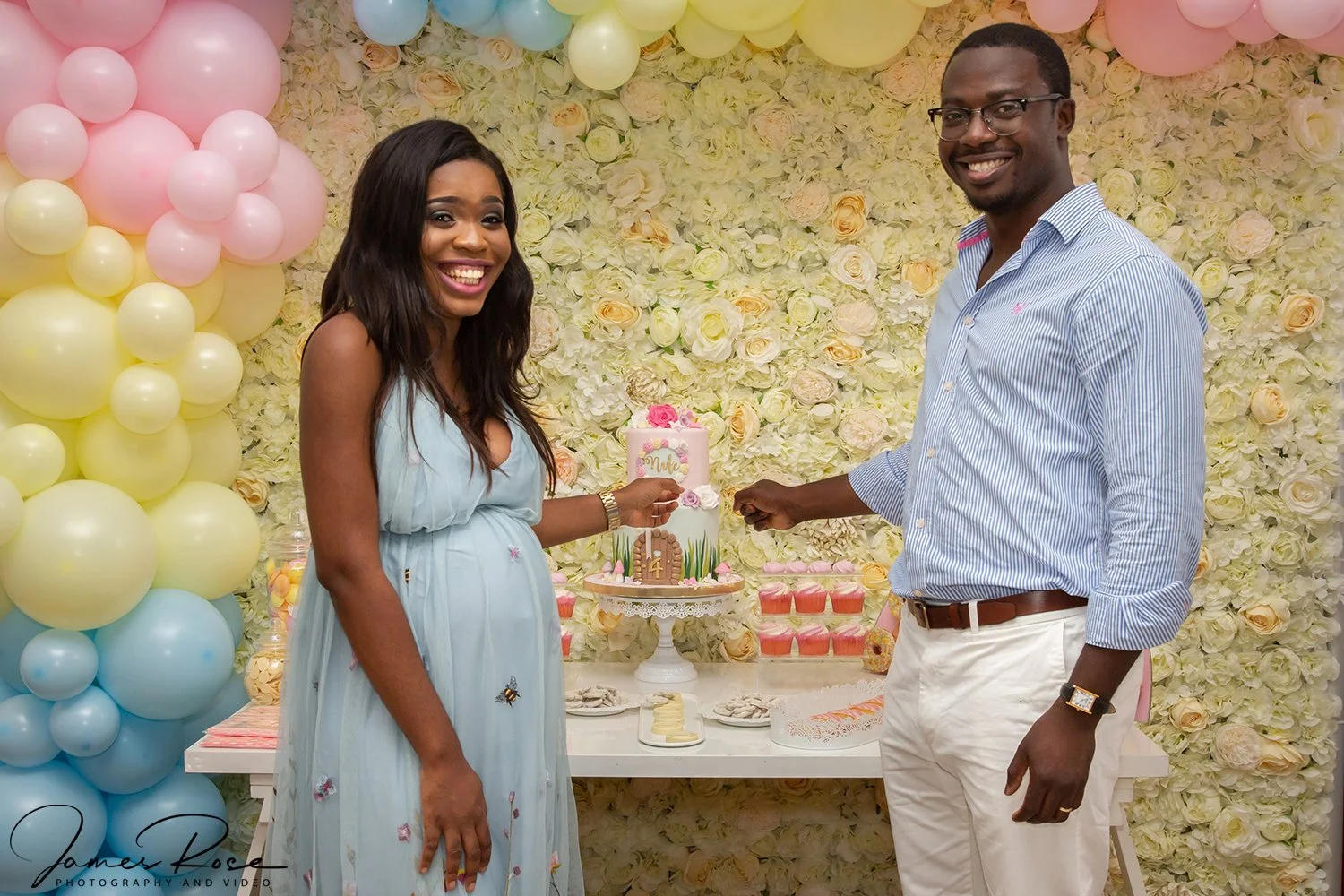 A woman and a man are celebrating at a party with a decorated floral and balloon backdrop. The woman is wearing a light blue dress, holding a slice of cake, and smiling. The man, dressed in a striped shirt and white pants, is holding the cake server.