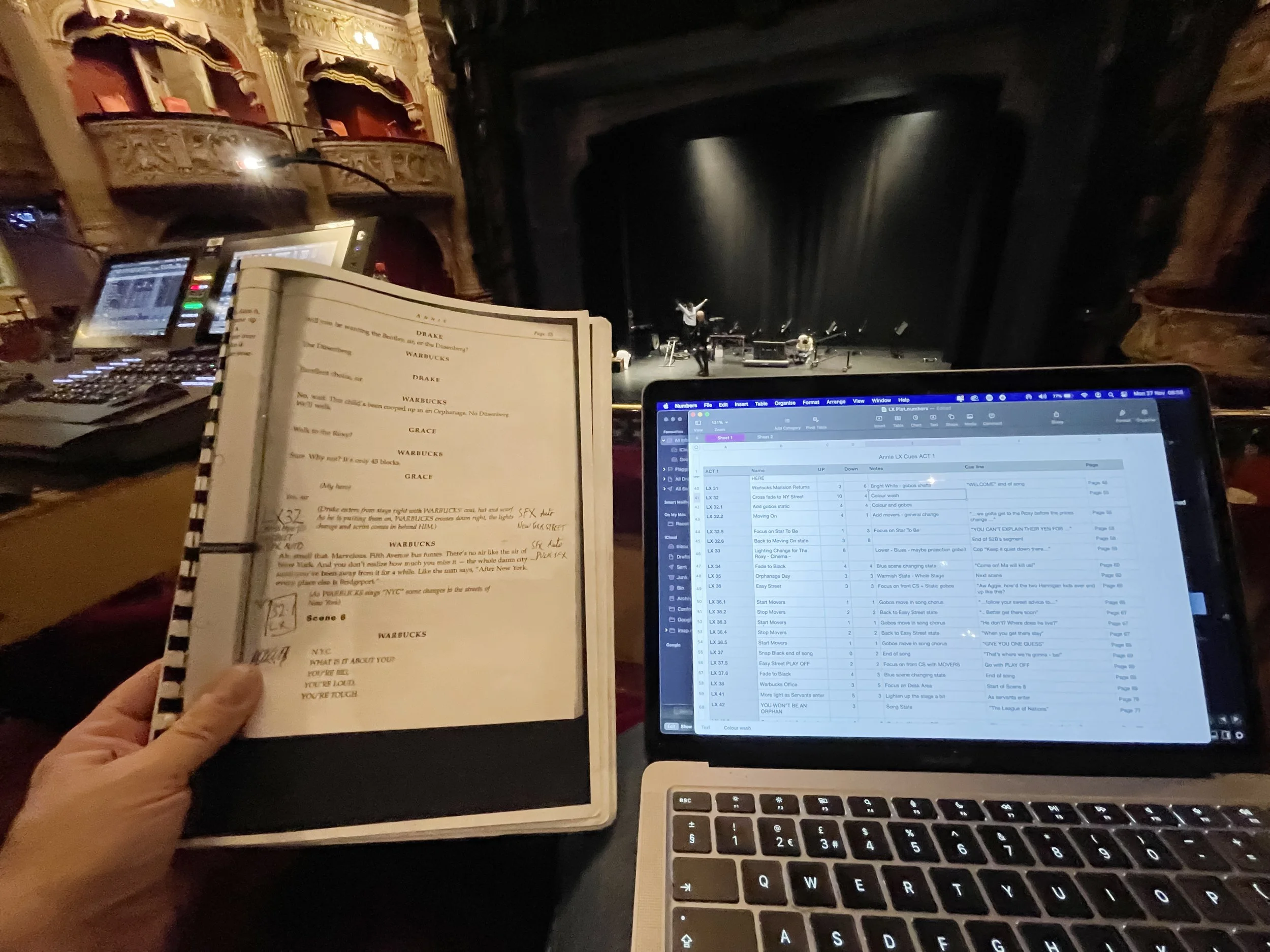 A person holding a script in one hand and operating a laptop with a screenplay and production notes on screen, while on a theater stage with a conductor and musicians rehearsing in the background.