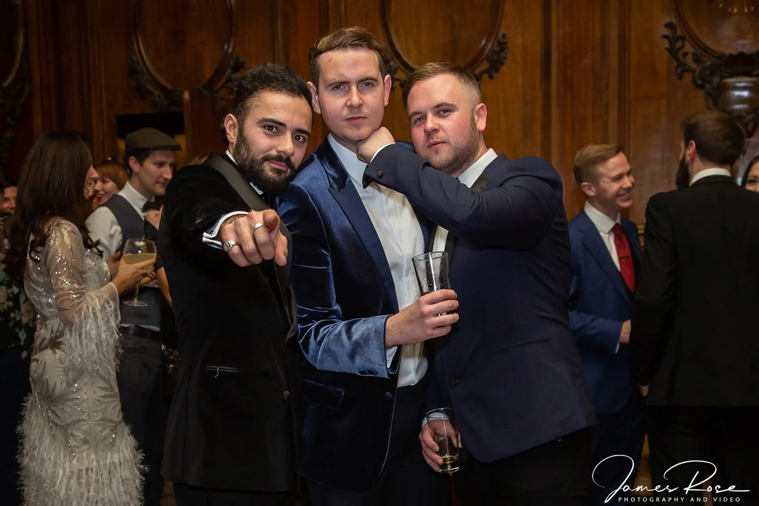 Three men in formal attire posing at a party or event, with a wood-paneled wall in the background. One man in the middle is wearing a blue velvet jacket, holding a drink, and the other two are interacting with him, one pointing at the camera and the 