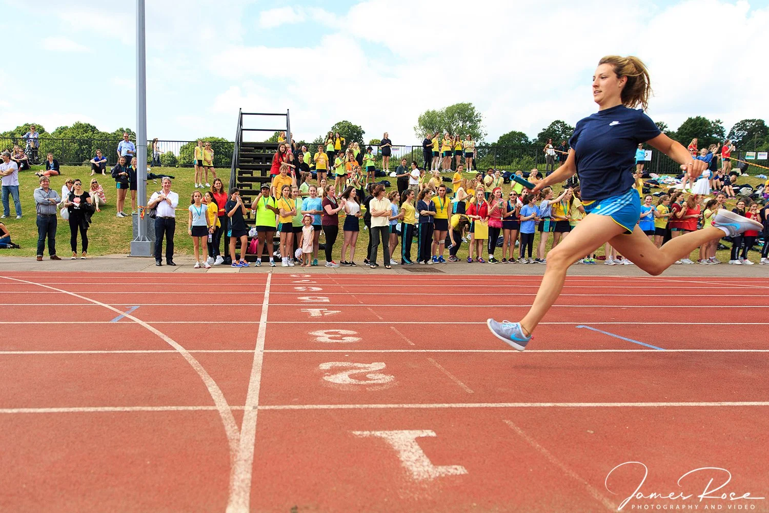 Female athlete in blue sportswear leaping in a track race with spectators watching on the sidelines.