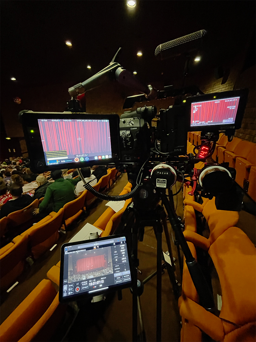 Camera setup on a tripod filming an indoor event with attendees seated and a red curtain stage in the background.