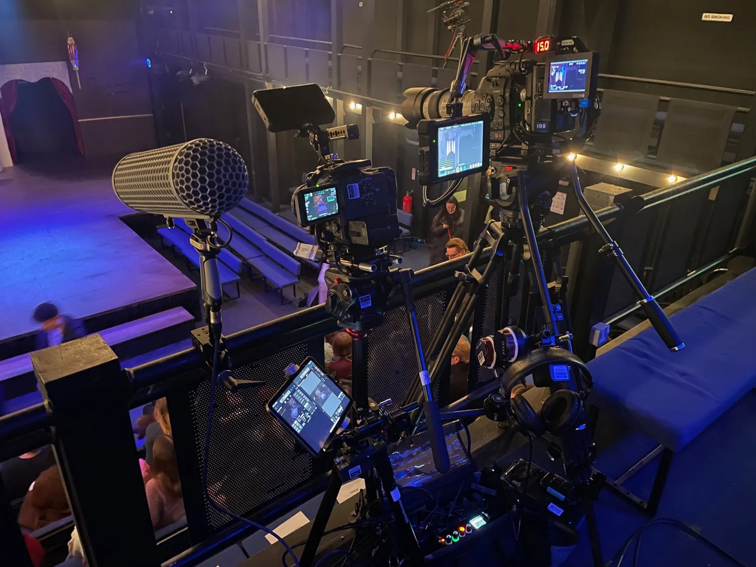 Cameras and filming equipment set up at an indoor venue with a stage and audience.