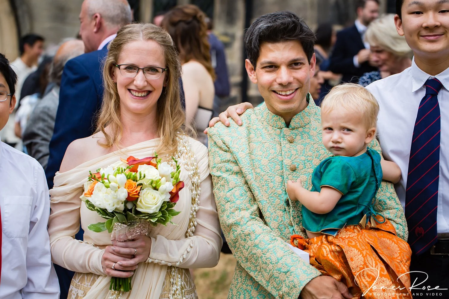 Smiling woman in cream dress holding a bouquet of colorful flowers, standing next to a smiling man in traditional teal and gold outfit holding a young child in green shirt, at an outdoor event with people in the background.