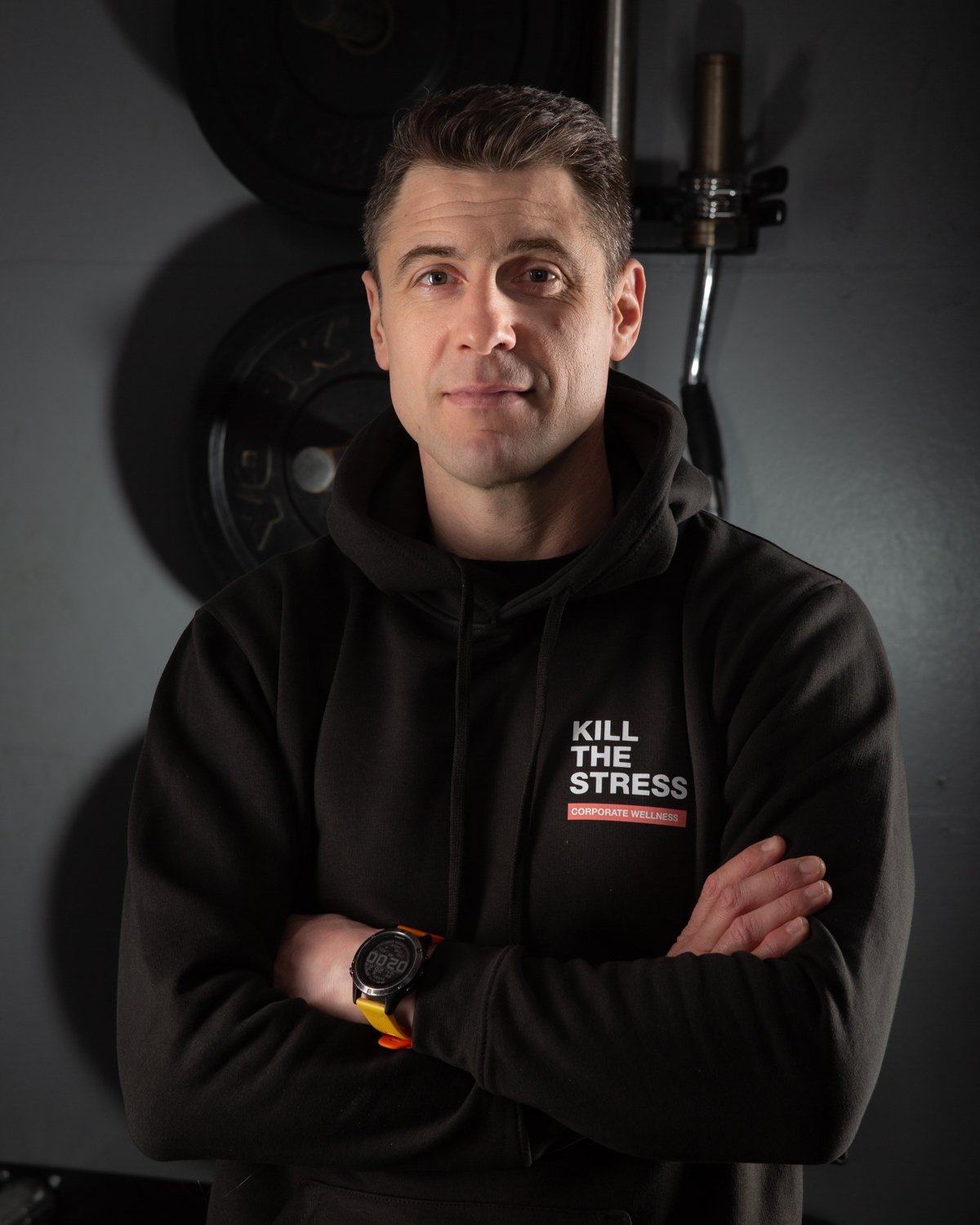 A man with short dark hair and fair skin, wearing a black hoodie with the phrase 'Kill the Stress' and 'Corporate Wellness', standing in a gym with weights in the background.