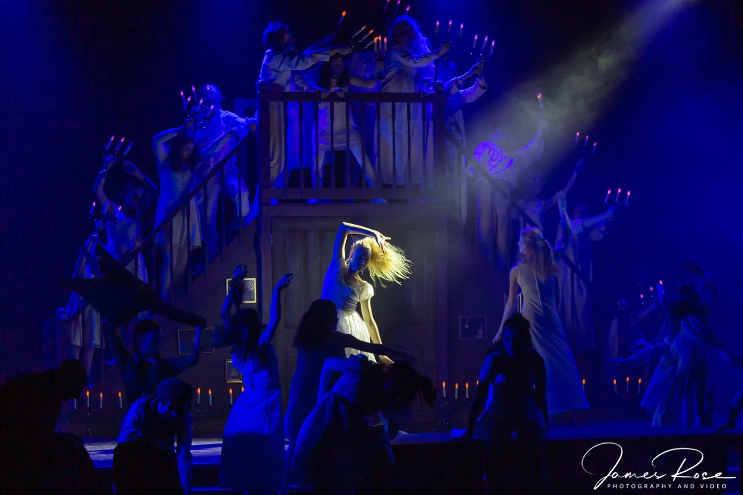 Scene from a theatrical dance performance with actresses in white dresses, some on stage and some on a balcony, under blue lighting, with candle-like lights in the background.
