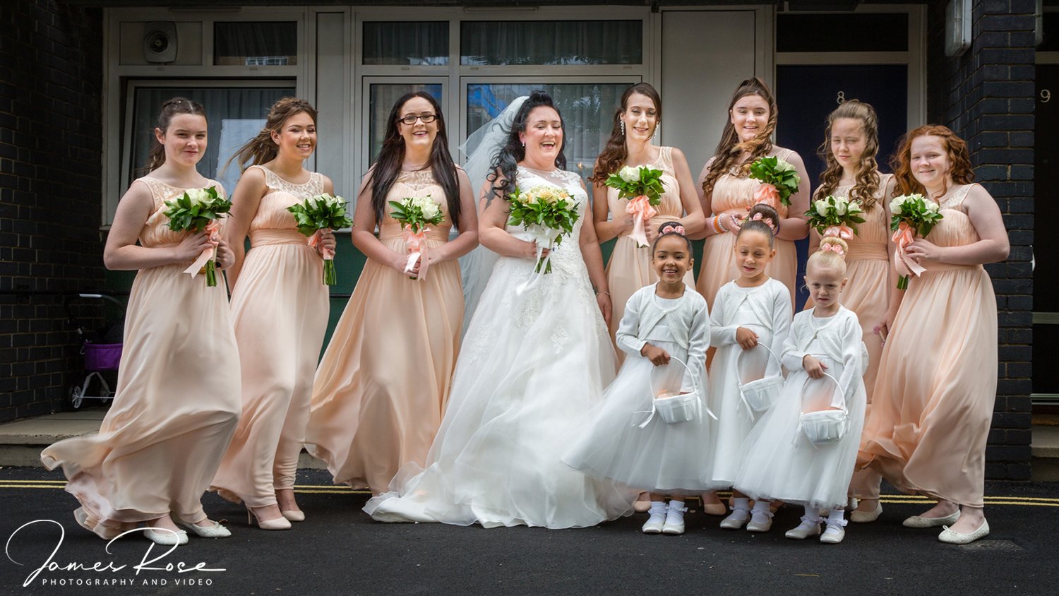 Group of women and young girls dressed in wedding attire standing outside a building, celebrating a wedding.
