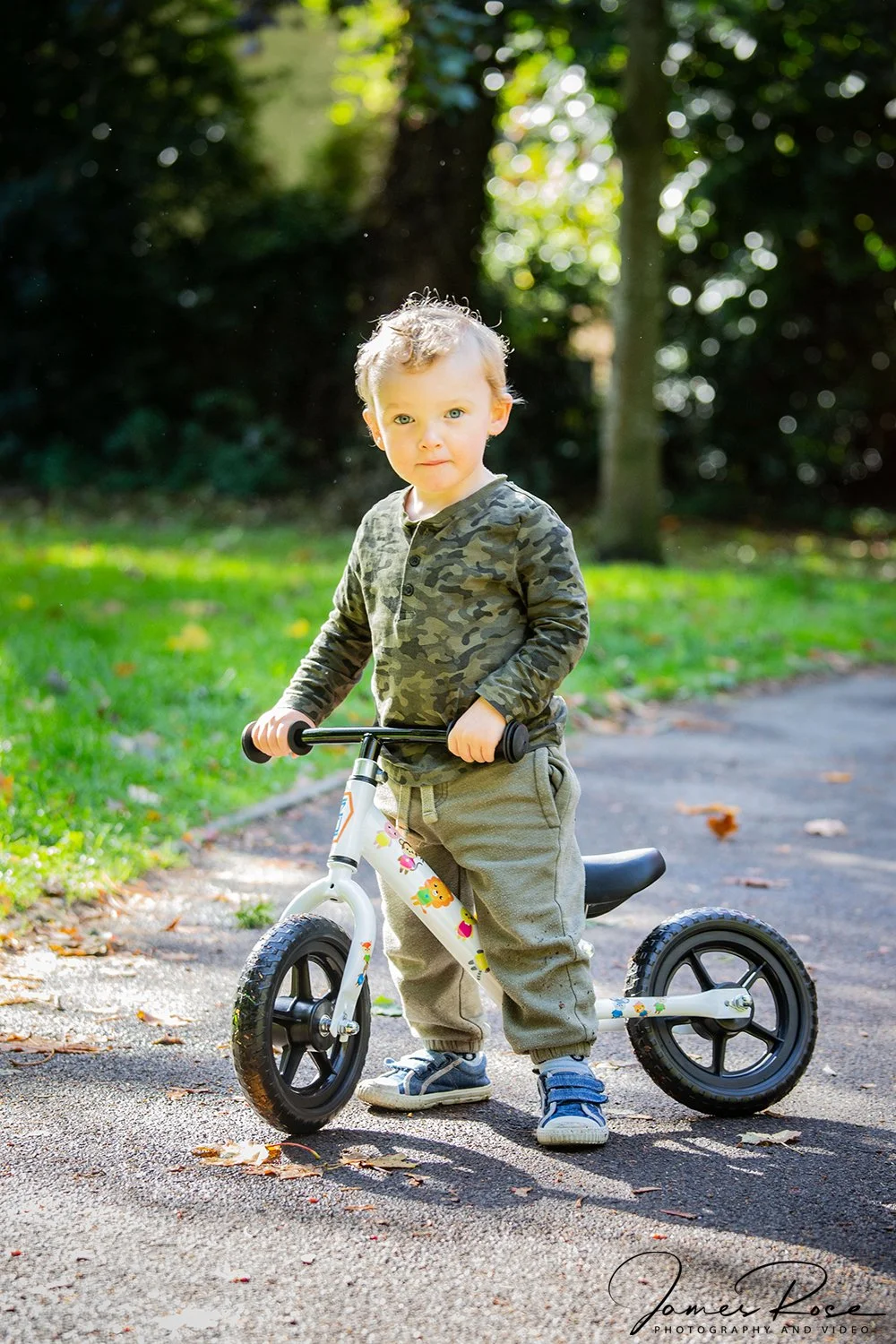 A young boy with curly hair and blue eyes standing next to a small white balance bike with colorful stickers, on a paved path in a park with green grass and trees in the background.