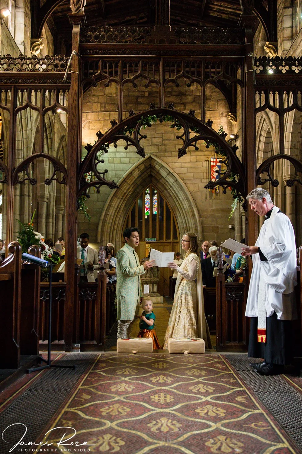 A wedding ceremony inside a church with a Gothic interior, where a couple is exchanging vows, accompanied by a minister, surrounded by guests.