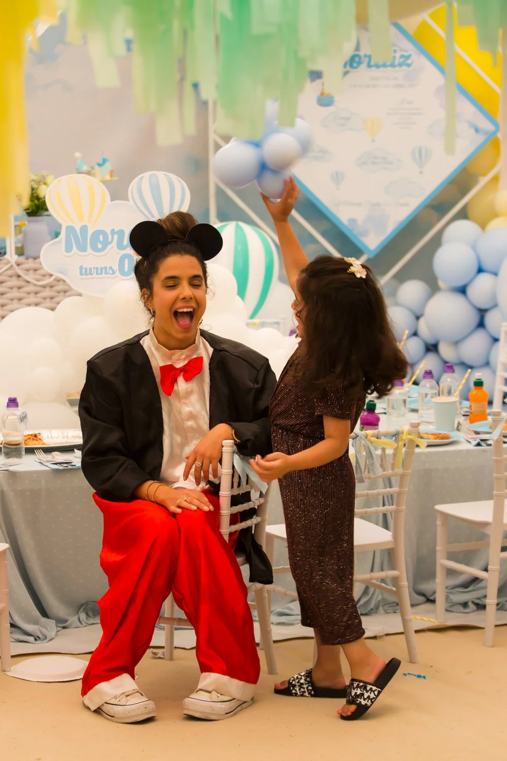 Two girls celebrating at a party with balloons and decorations. One dressed as Mickey Mouse with a black jacket, red pants, white shirt, and Mickey Mouse ears, sitting on a chair. The other girl in a black dress with floral hair clip and black slide 