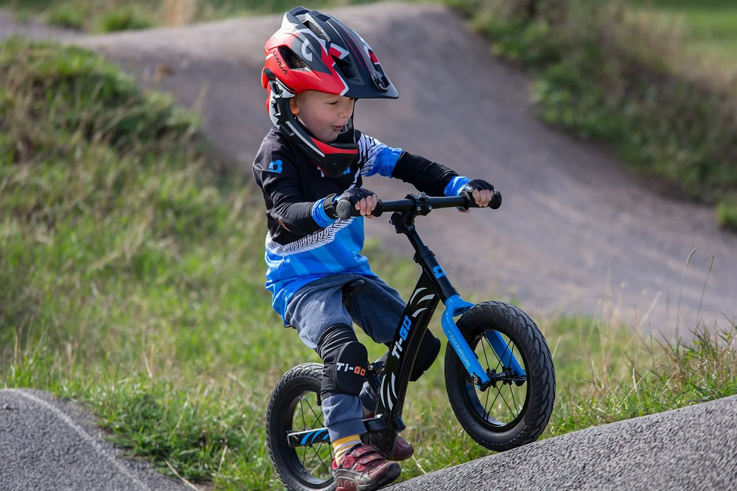 Young boy riding a balance bike on a dirt trail with grassy surroundings, wearing a helmet and protective gear.