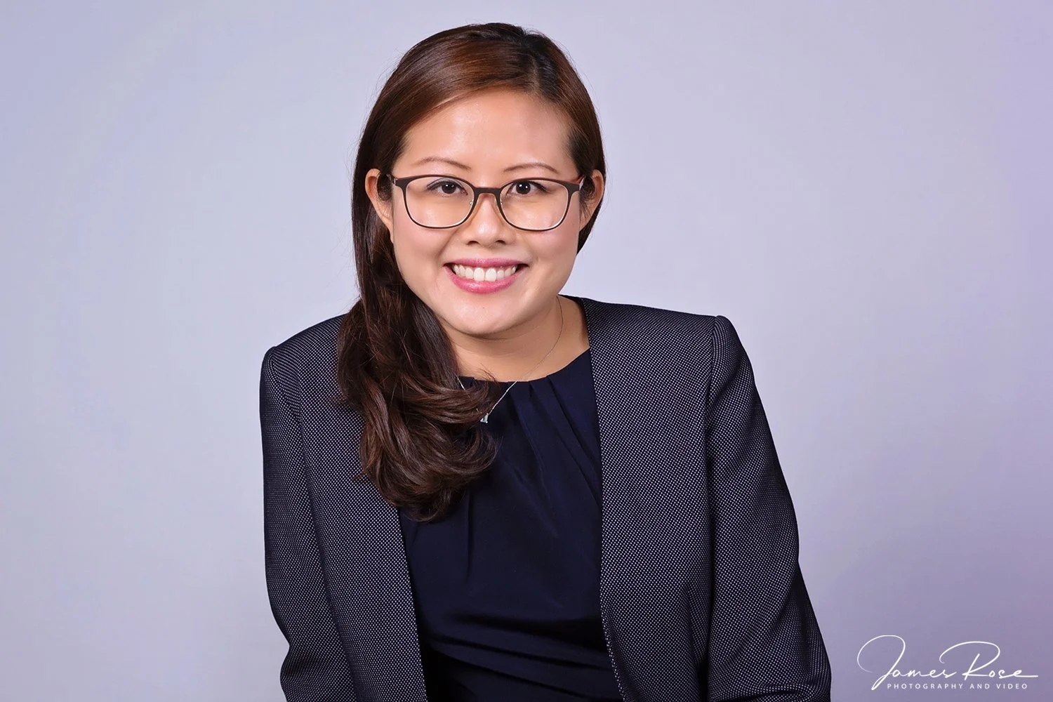 A woman with long brown hair, glasses, and a bright smile, wearing a black blazer and a navy blue top, posing against a plain light gray background.