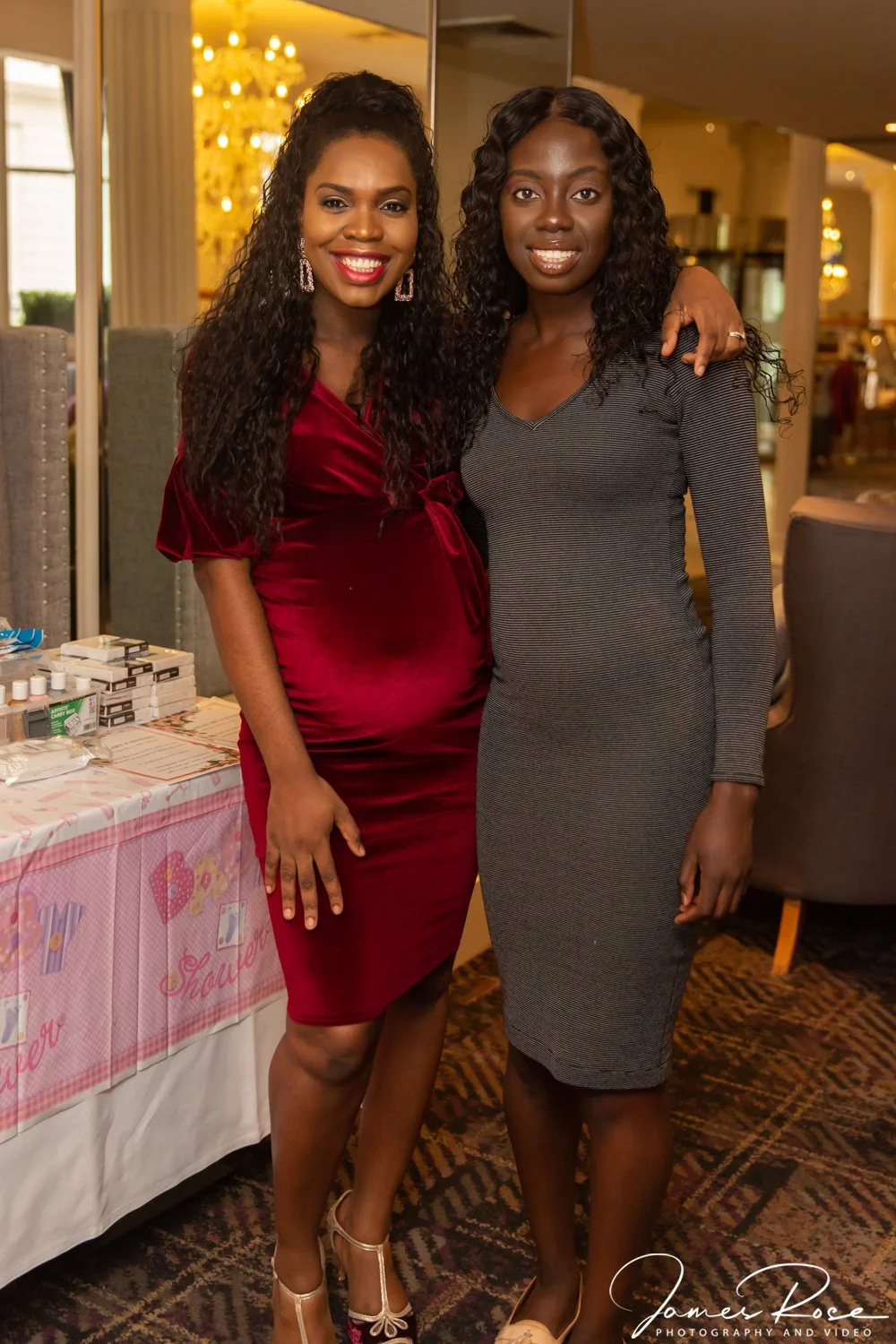 Two women standing together in an indoor setting, smiling at the camera. One is wearing a red velvet dress and the other a gray fitted dress. There are tables with various items and a pink-themed setup in the background.