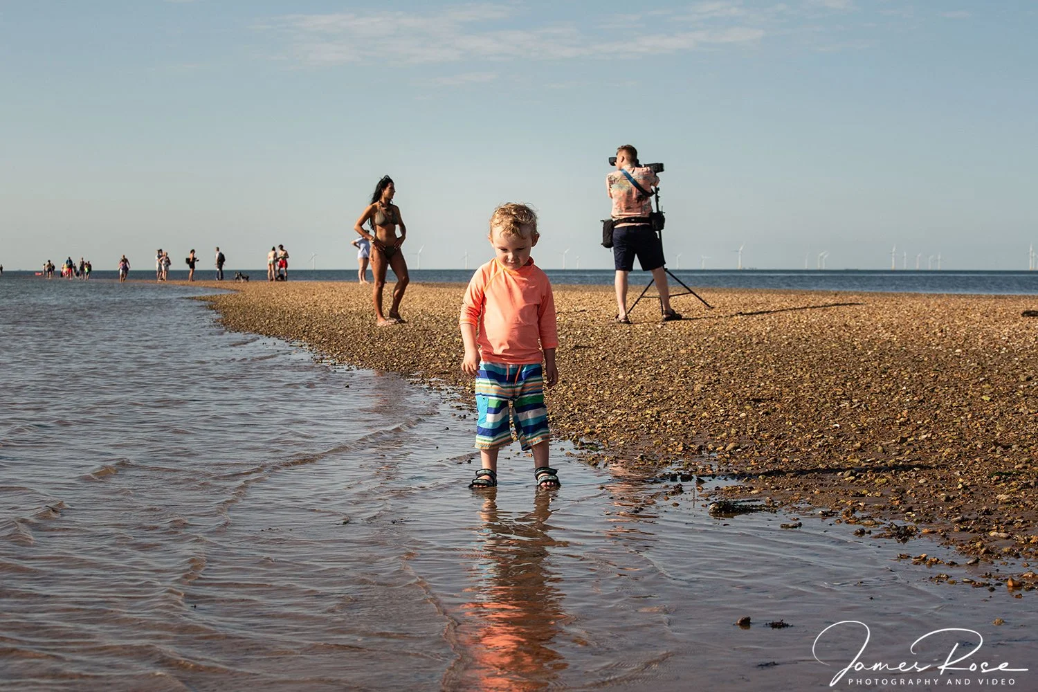 A young boy standing in shallow water at the beach with people in the background, including a woman in a swimsuit, a person operating a camera on a tripod, and others walking along the shoreline.