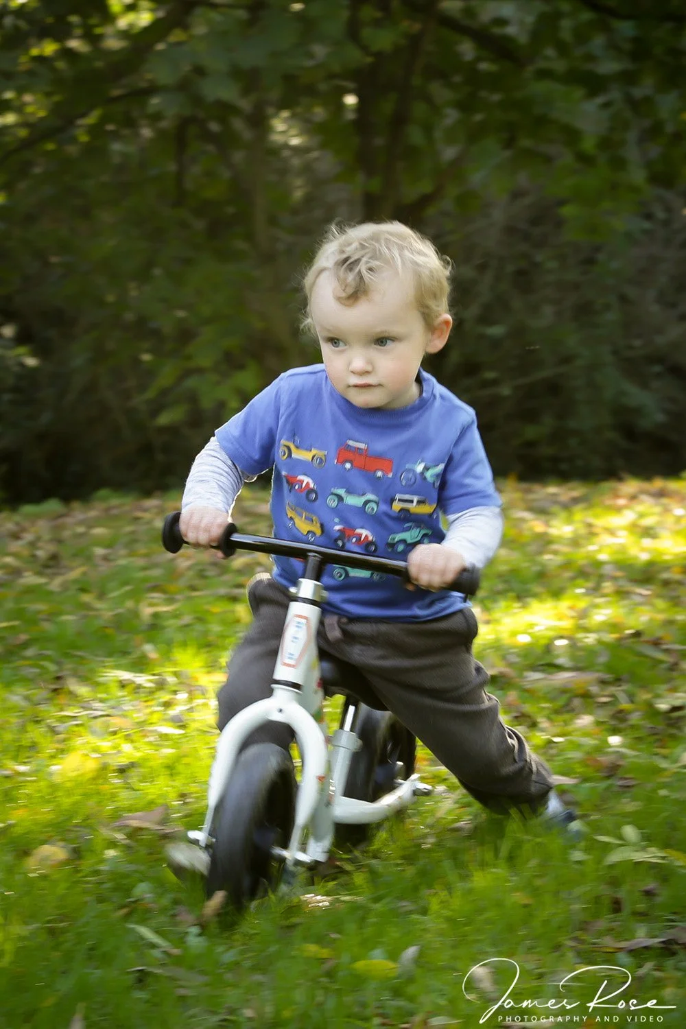 Young boy riding a bicycle on grass in a wooded area.