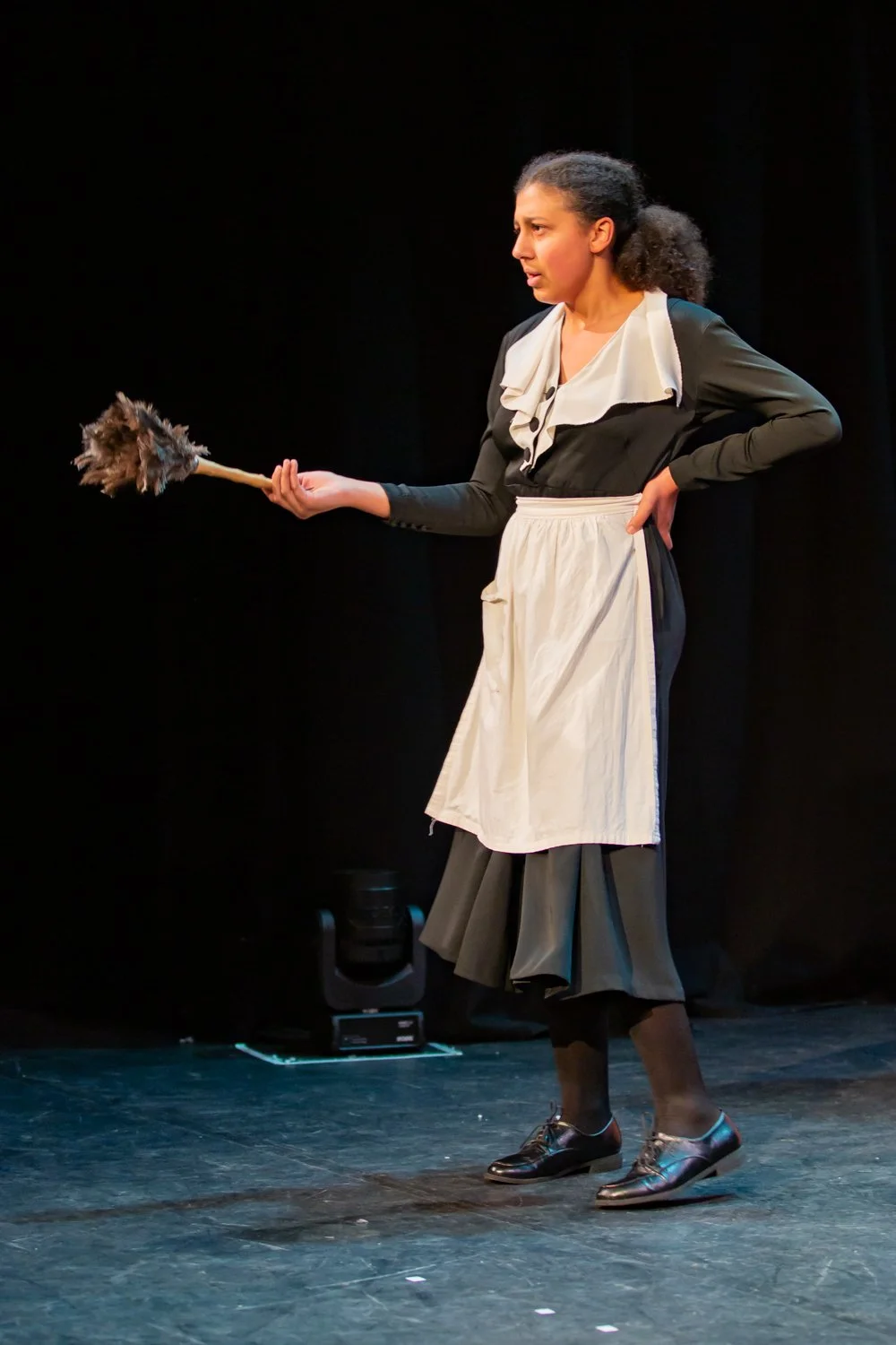 A woman dressed as a maid holding a feather duster on stage, with a black curtain background.