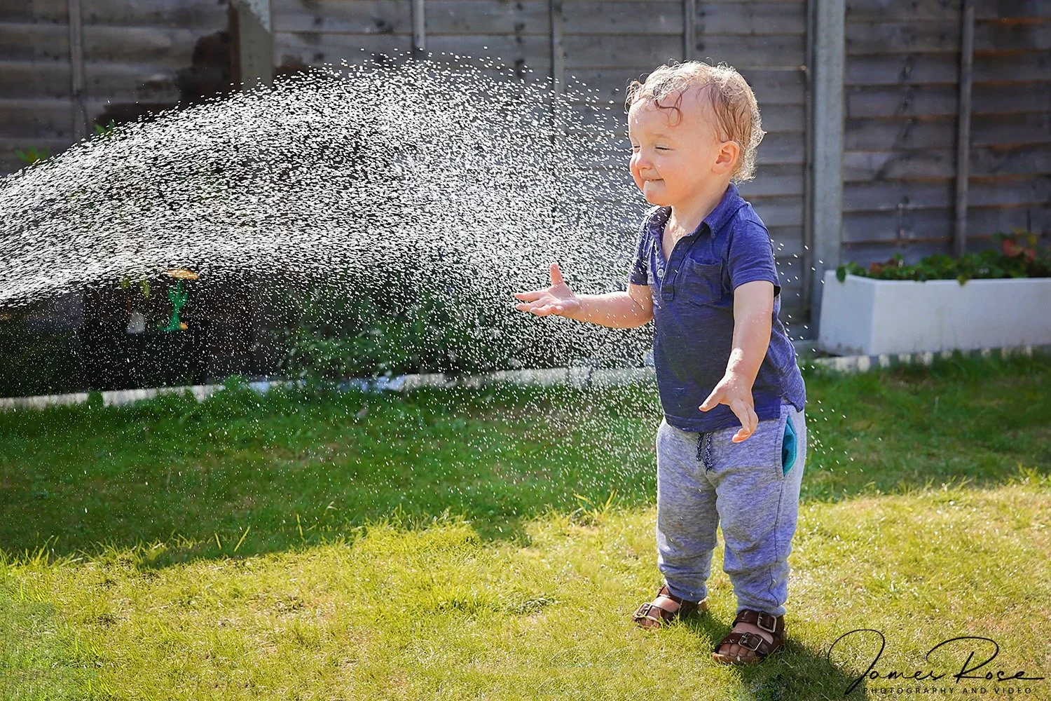 A young boy playing with water from a garden hose in a backyard. He is wearing a blue shirt, gray pants, and sandals, smiling as water splashes around him. Wooden fence and garden bed in the background.