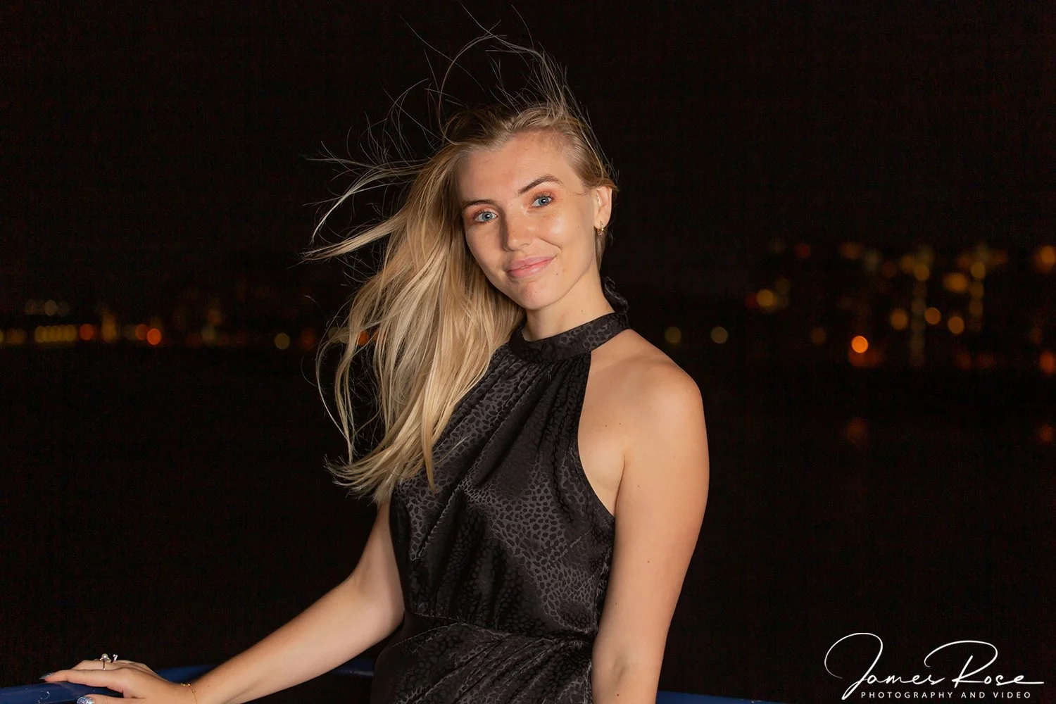 A young woman with long blonde hair, wearing a black sleeveless dress, stands outdoors at night with a city skyline in the background.