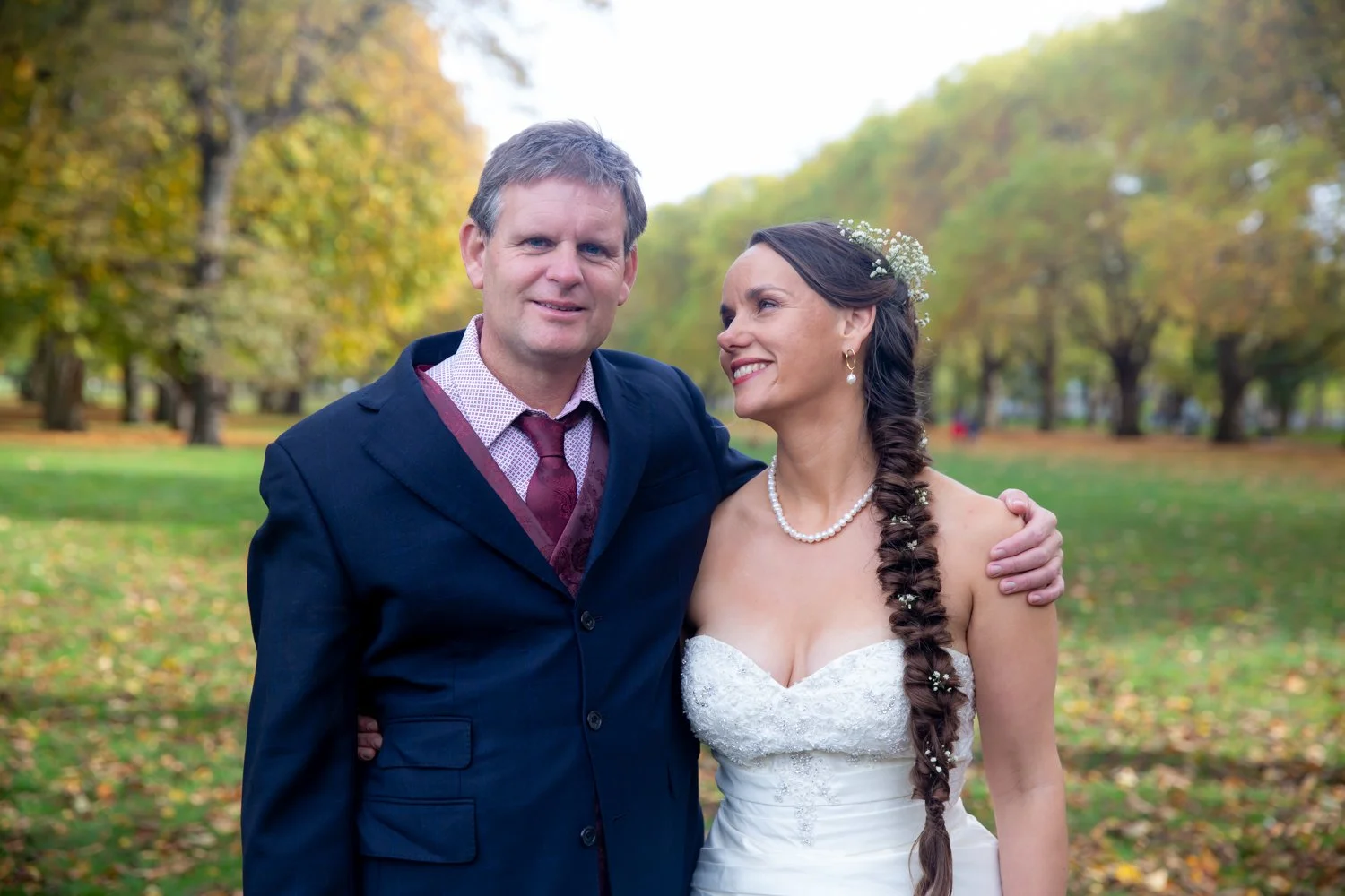 A man and a woman, dressed in a suit and wedding gown respectively, standing close together outdoors in a park with autumn trees in the background.