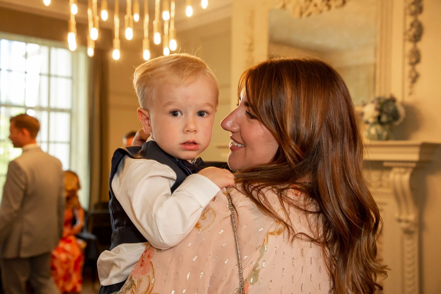 A woman with long reddish-brown hair smiling while holding a young blond boy in a fancy suit in an elegant room with warm lighting and chandeliers.