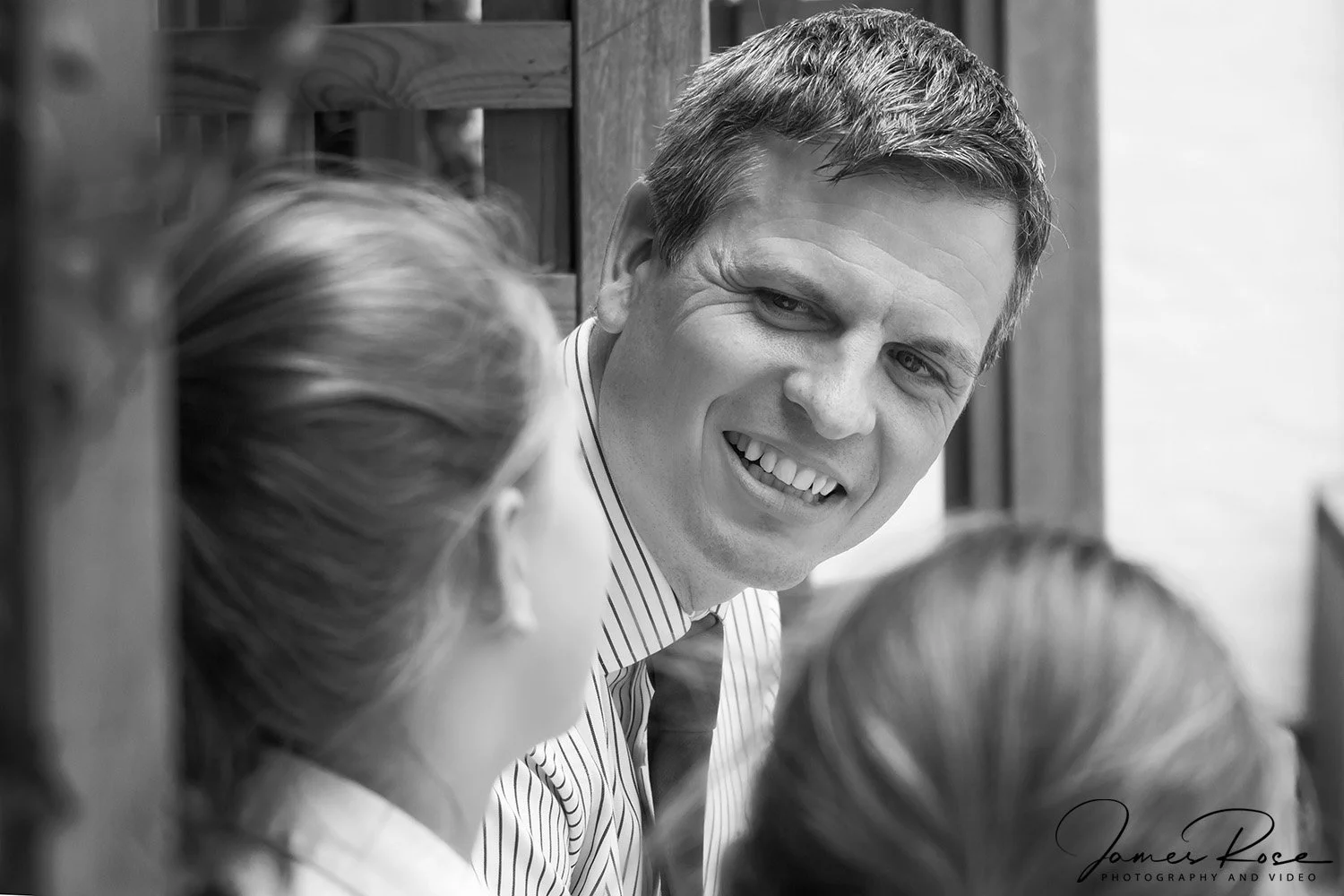 A black and white photo of a man smiling and looking at two women whose faces are partly visible. The man has short hair and is wearing a striped shirt and tie.