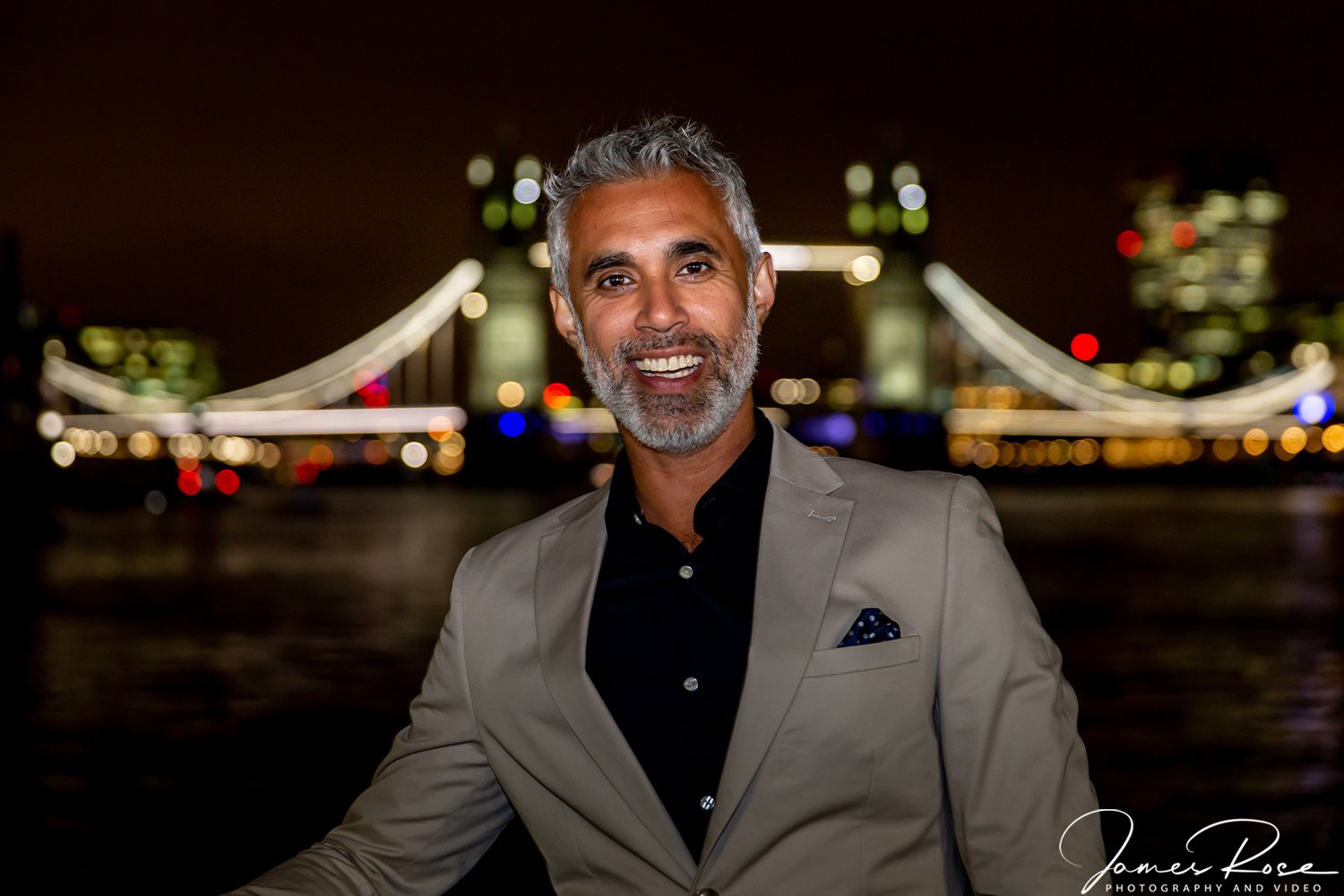 A smiling man with gray hair and a beard, wearing a beige blazer and black shirt, standing outdoors at night with the illuminated Tower Bridge in London in the background.