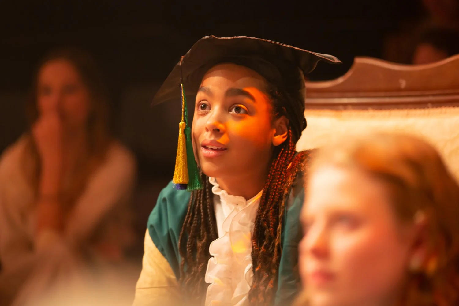 A woman in a graduation cap and gown talking at a graduation ceremony, with others listening in the background.