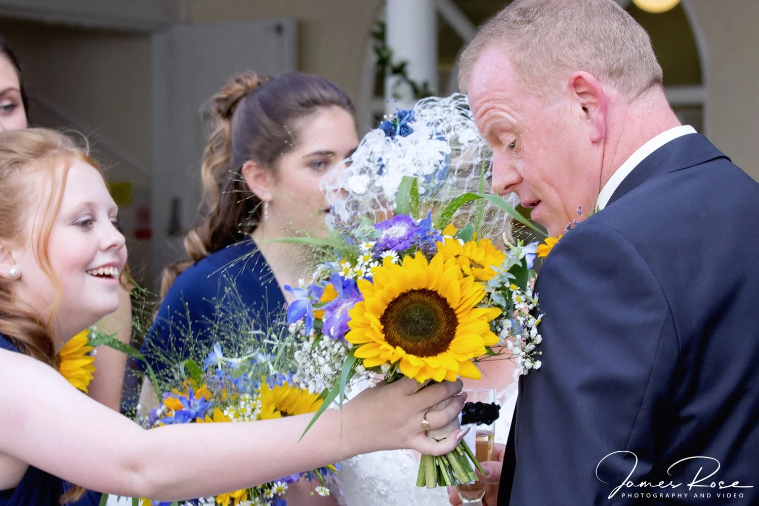 A man in a suit receives a large bouquet of sunflowers and other flowers from a smiling red-haired woman at a wedding or celebration event.