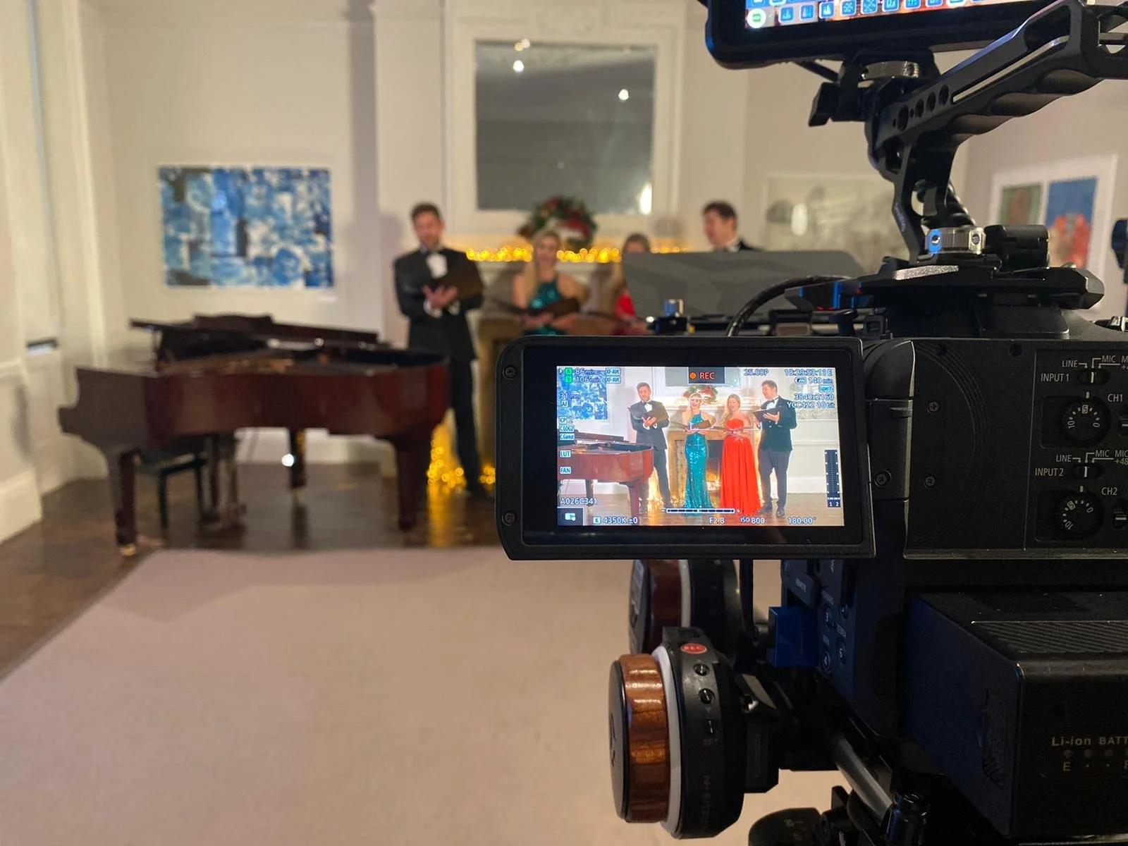 A video camera recording a performance of four people dressed in formal attire on a stage with a grand piano. The scene is viewed on the camera's display screen.