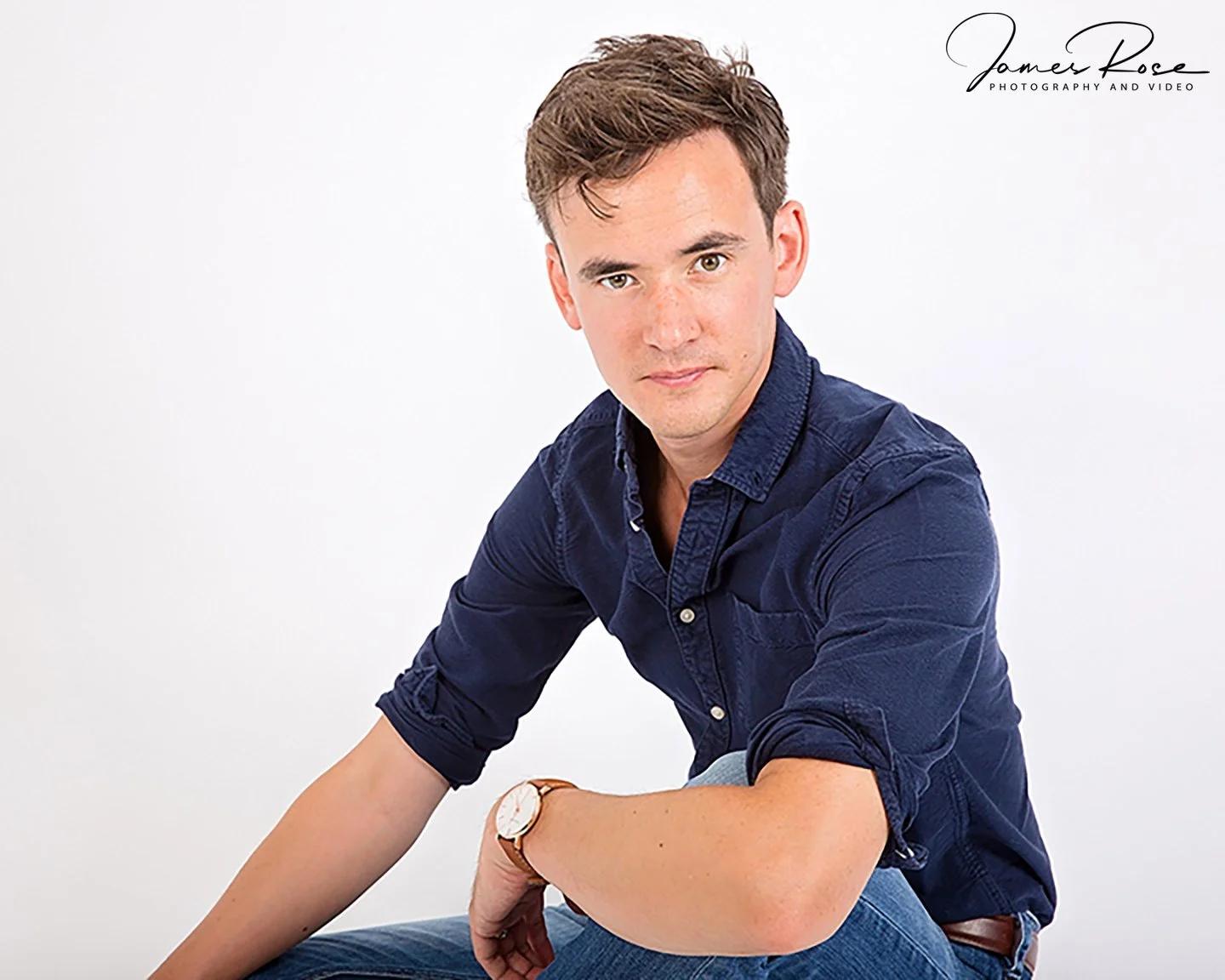 A young man with brown hair sitting against a white background, wearing a navy blue shirt and blue jeans, with a wristwatch on his left wrist and looking at the camera.