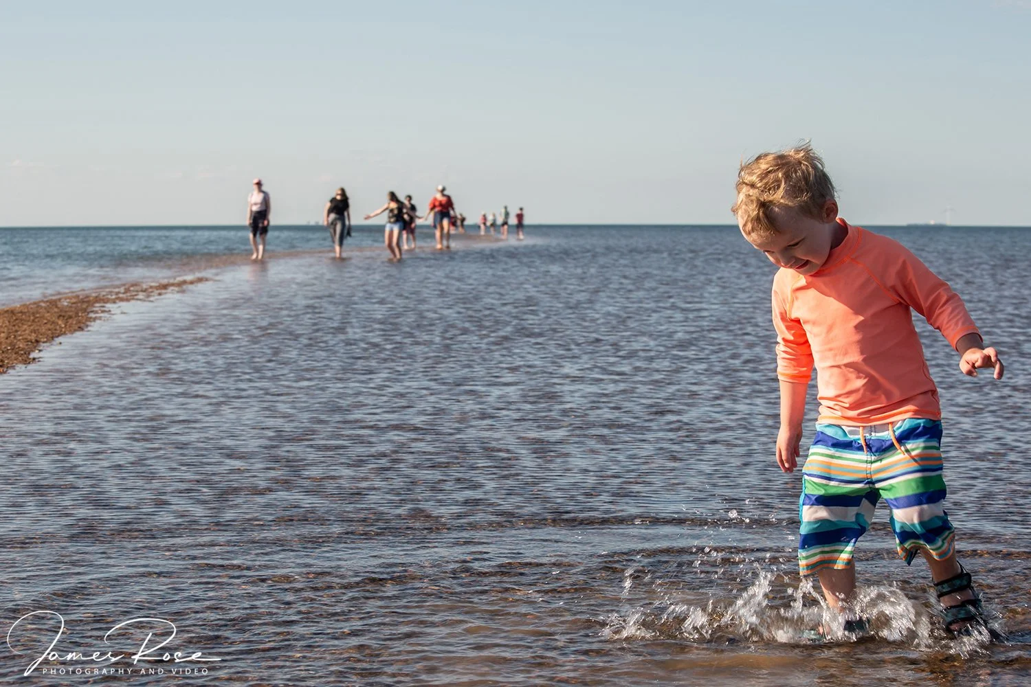 A young boy in colorful striped shorts and an orange shirt playing in shallow water at the beach with a group of people walking in the distance.