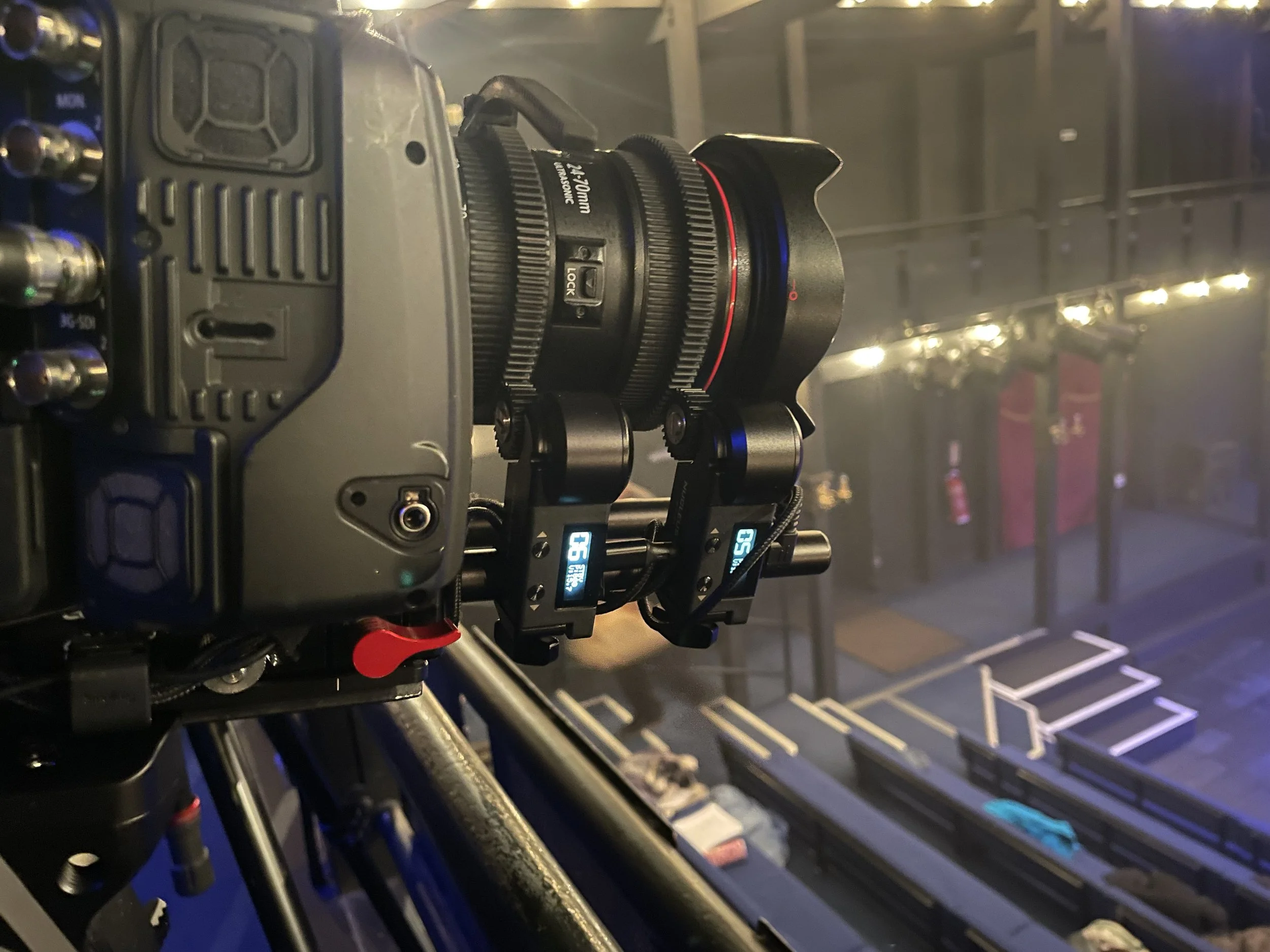 Close-up of a professional camera mounted on a stabilizer, facing towards a theater stage with set pieces and lighting.