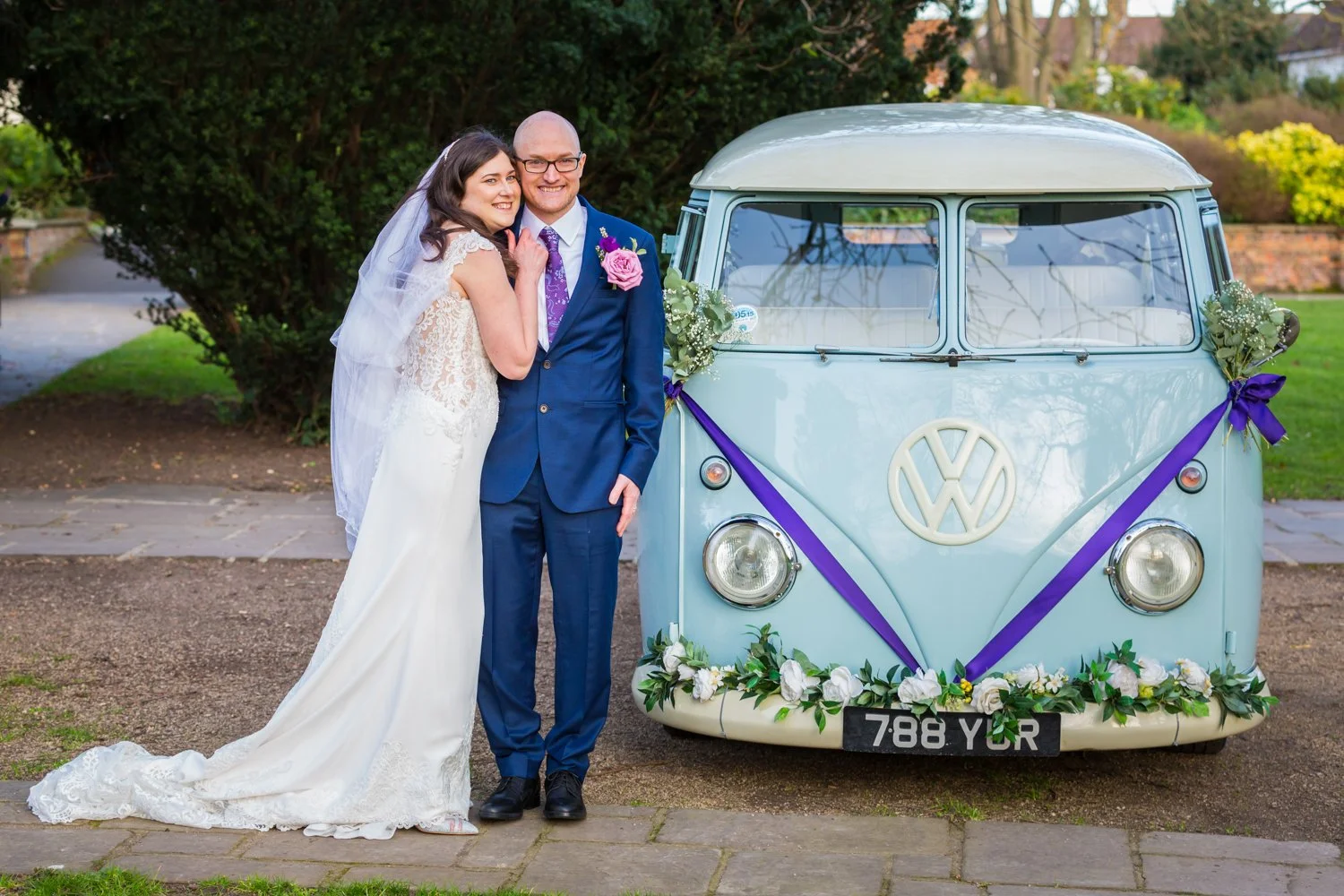 A bride and groom in wedding attire standing beside a vintage light blue Volkswagen van decorated with flowers and ribbons. The bride wears a white lace wedding gown with a veil, and the groom wears a navy suit with a purple tie and pink boutonniere.