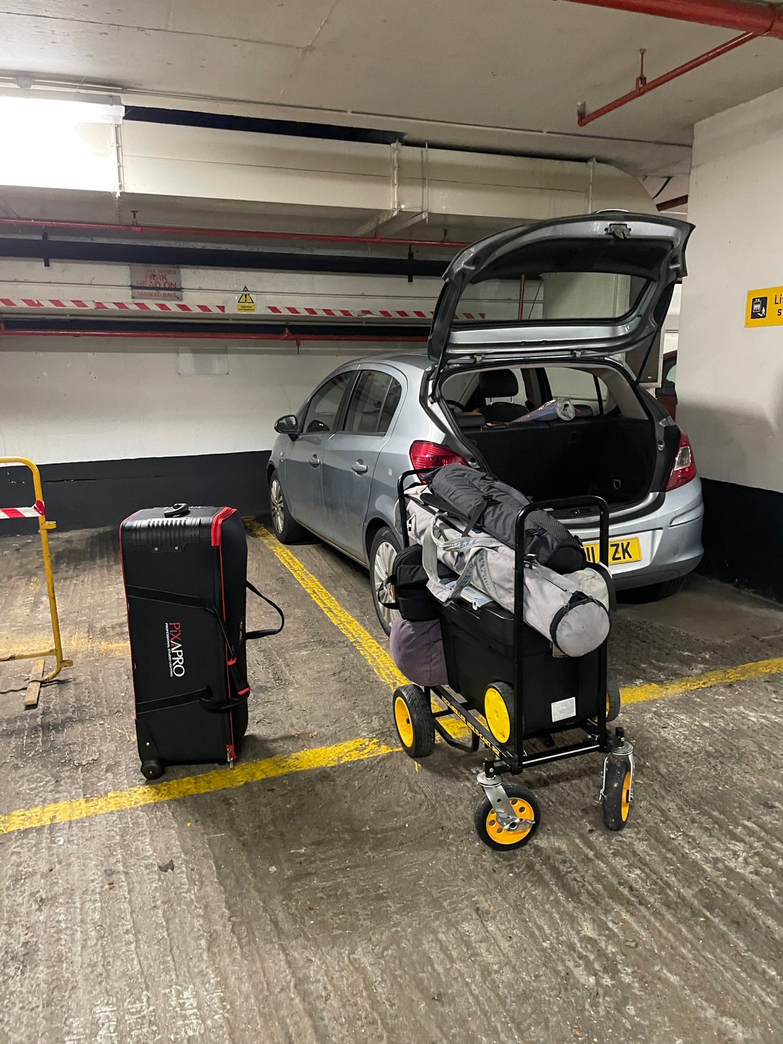 A parking garage with a silver hatchback car, an open trunk, a black suitcase, and a luggage cart filled with bags and gear.