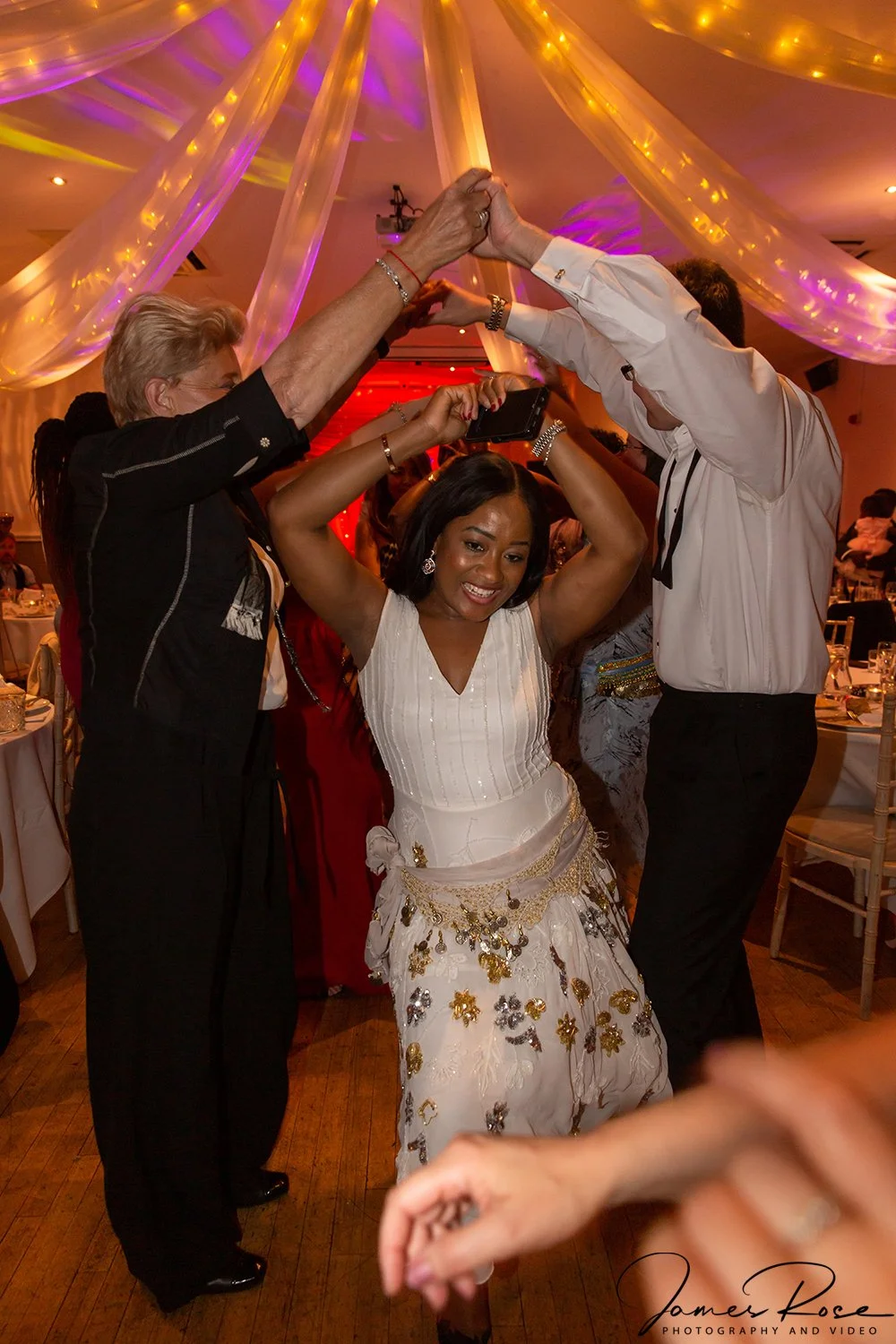 A woman wearing a white dress decorated with gold and silver embellishments is dancing with two men, one on each side, at a lively event with colorful lighting and draped fabric decorations.