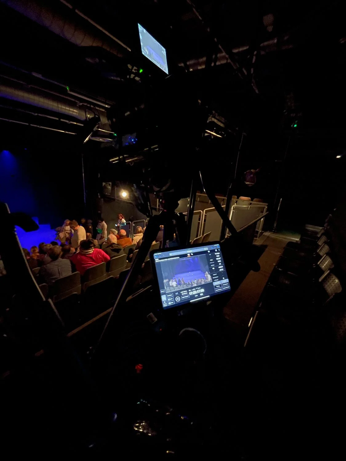 Inside a dark theater with a camera operator in the foreground, filming an audience seated under blue stage lighting.