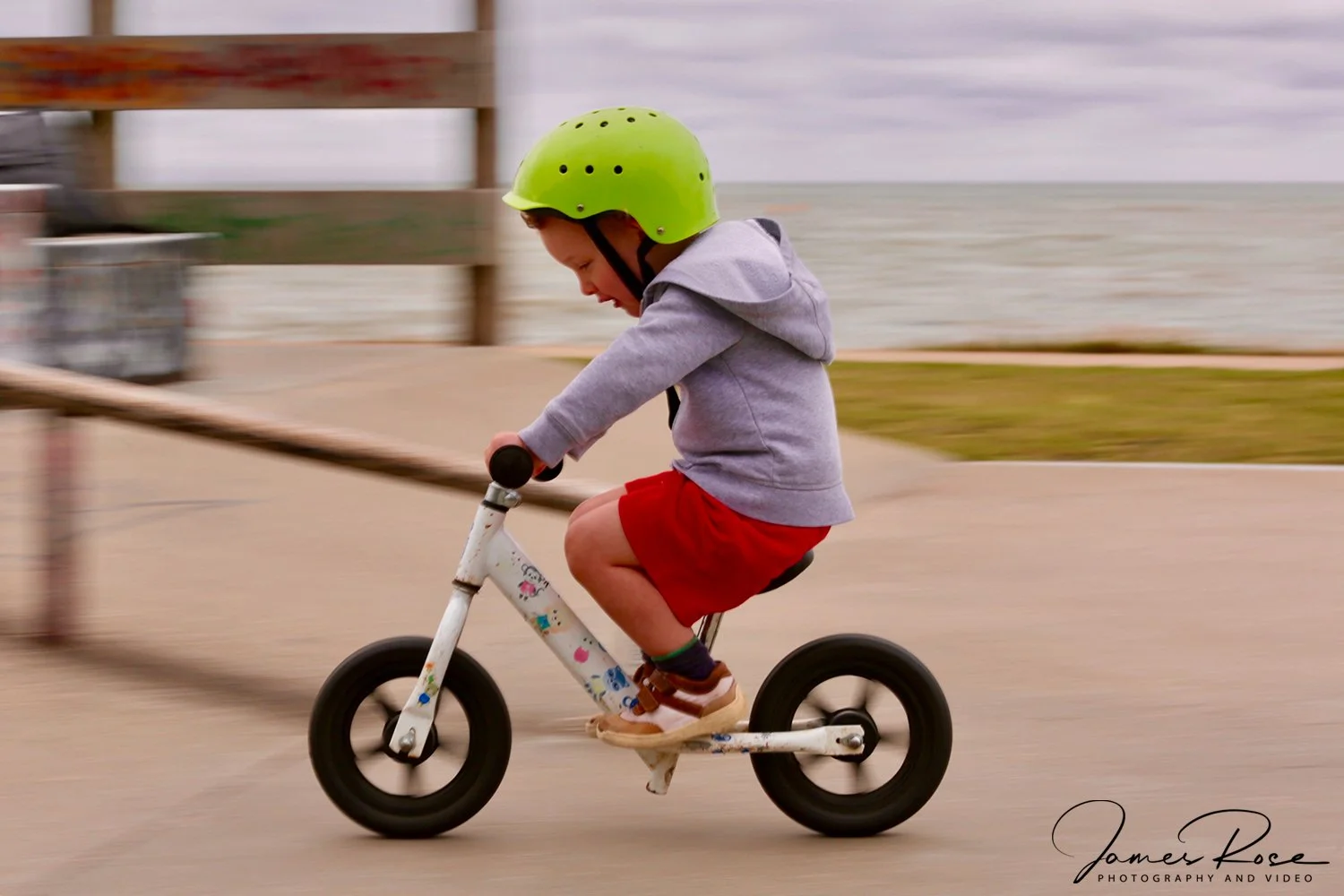A young boy wearing a green helmet, gray hoodie, and red shorts riding a small white bicycle along a paved path near the water.