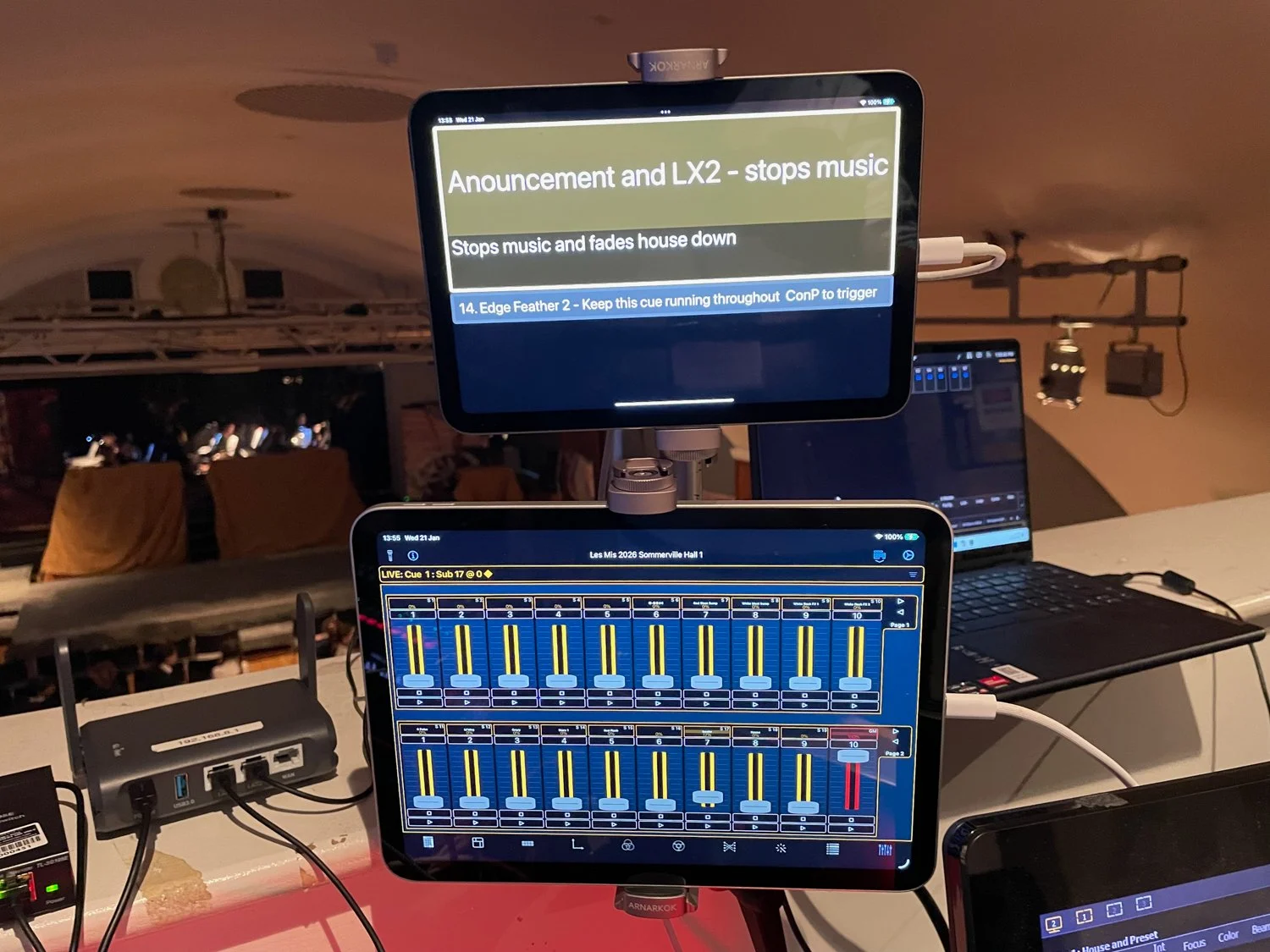 Photo of a live music venue control setup with a tablet and monitor showing sound cue information, a laptop, and various audio equipment in the background.