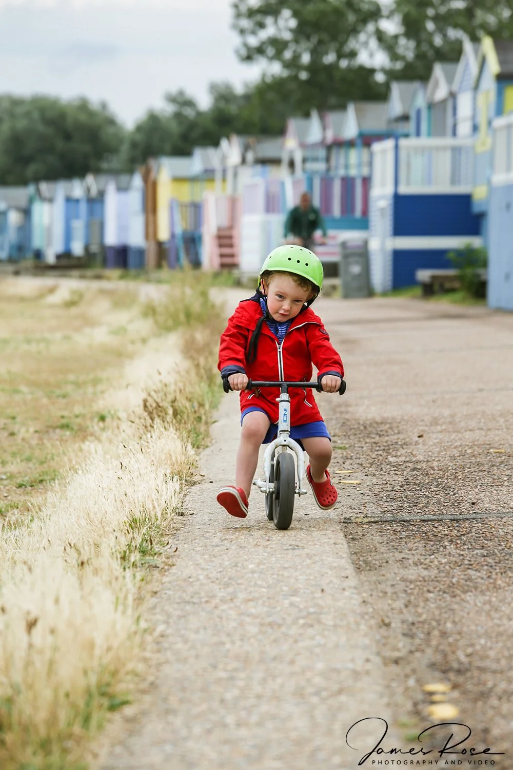 A young boy wearing a green helmet, red jacket, and red Crocs riding a balance bike on a pathway with colorful beach huts in the background.