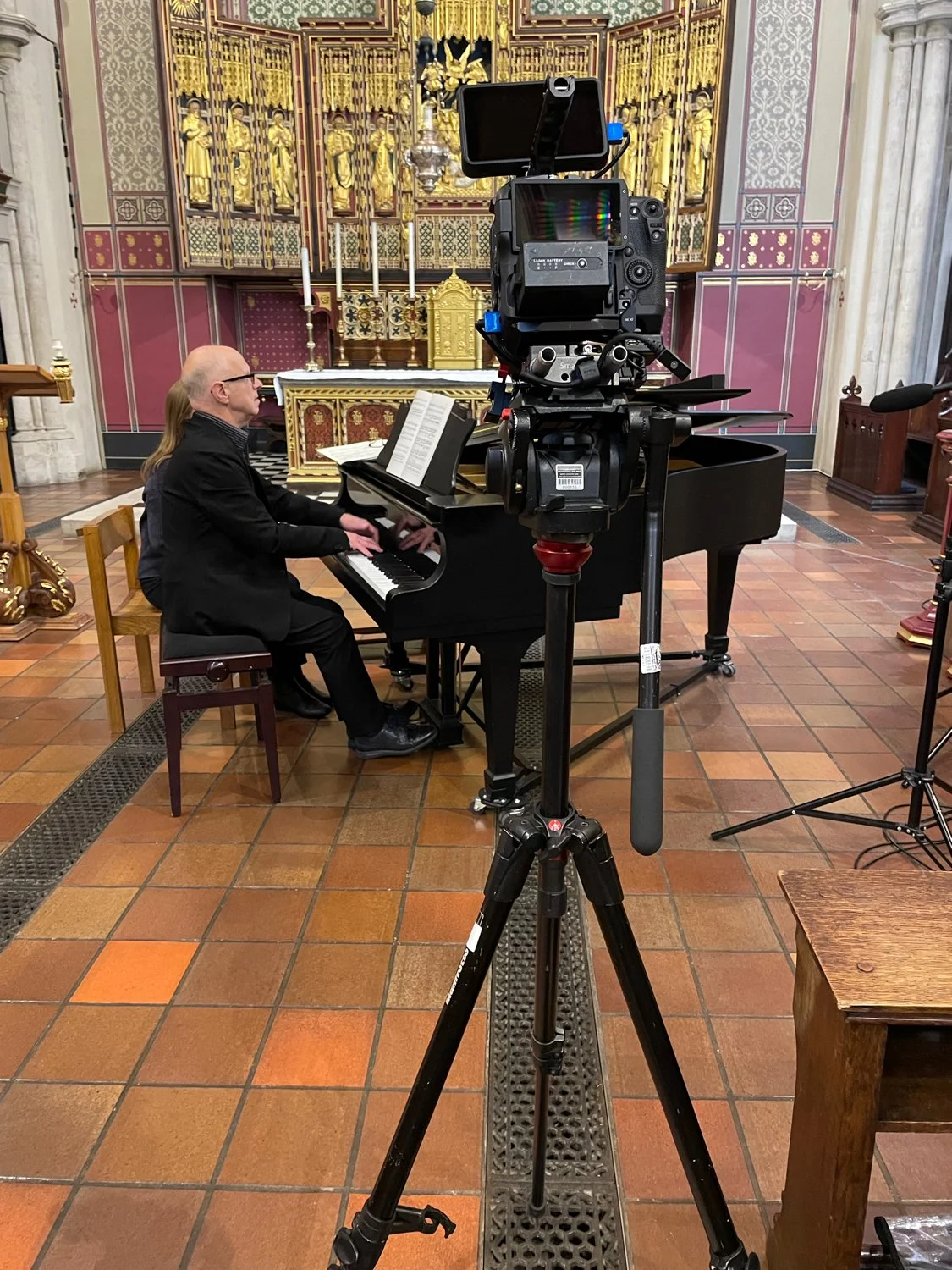 A man playing a grand piano in a church or chapel with ornate gold detailing and religious statues in the background. A camera on a tripod is filming the performance.